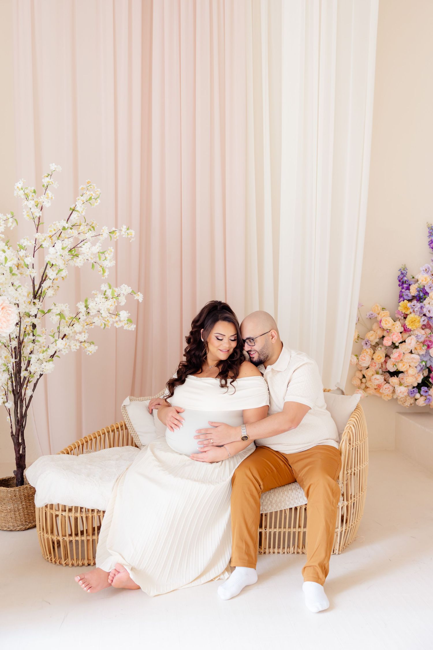 A couple sits together on a rattan bench surrounded by floral arrangements in a pink and white studio setting.