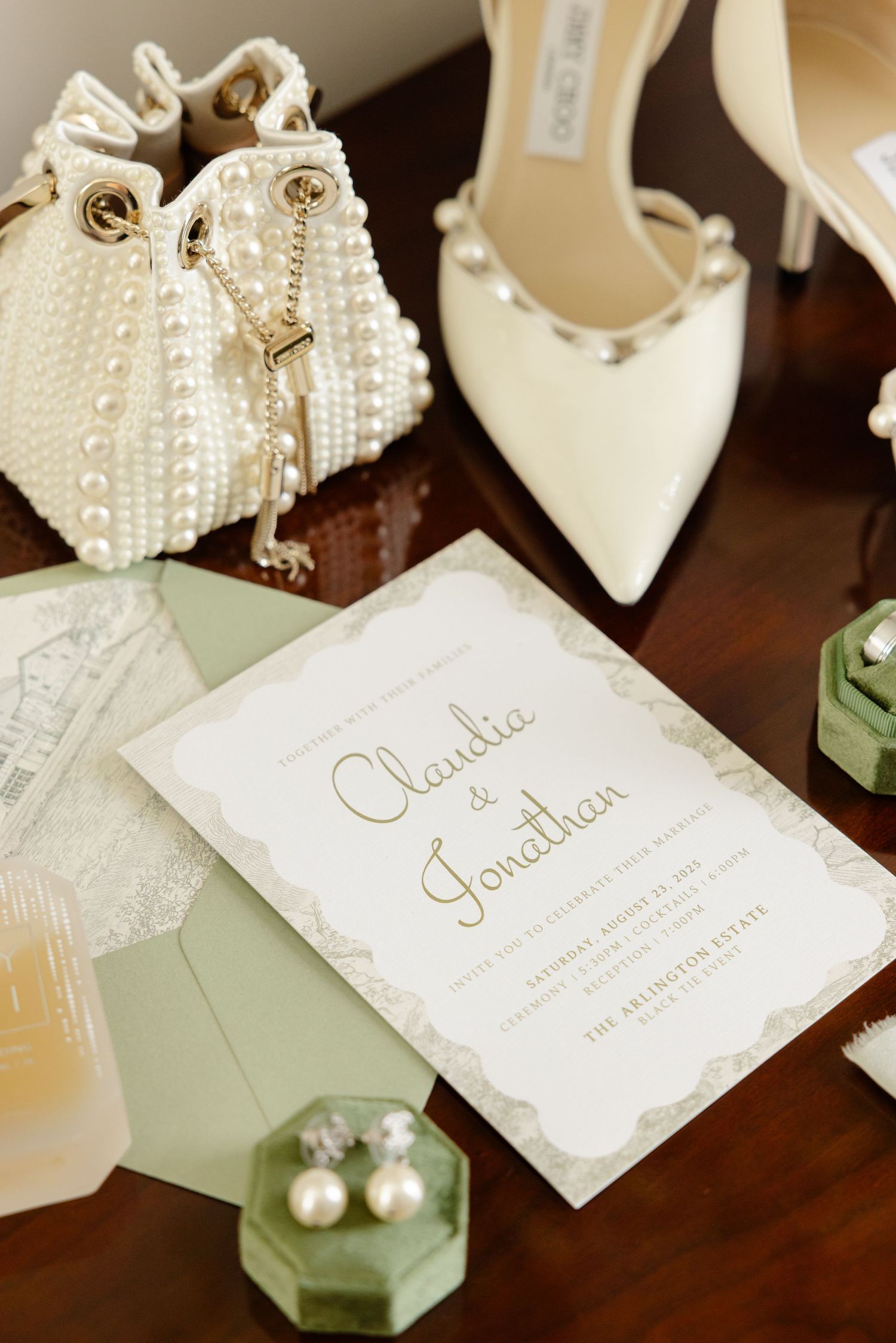 Elegant wedding invitation with pearl-adorned shoes and white beaded purse on wooden surface.