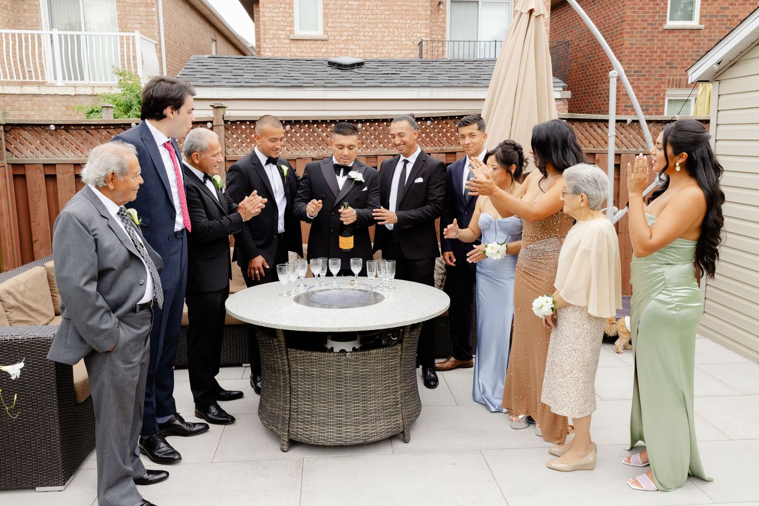 Outdoor wedding reception guests gather around a circular fire pit table on a brick patio during evening festivities.