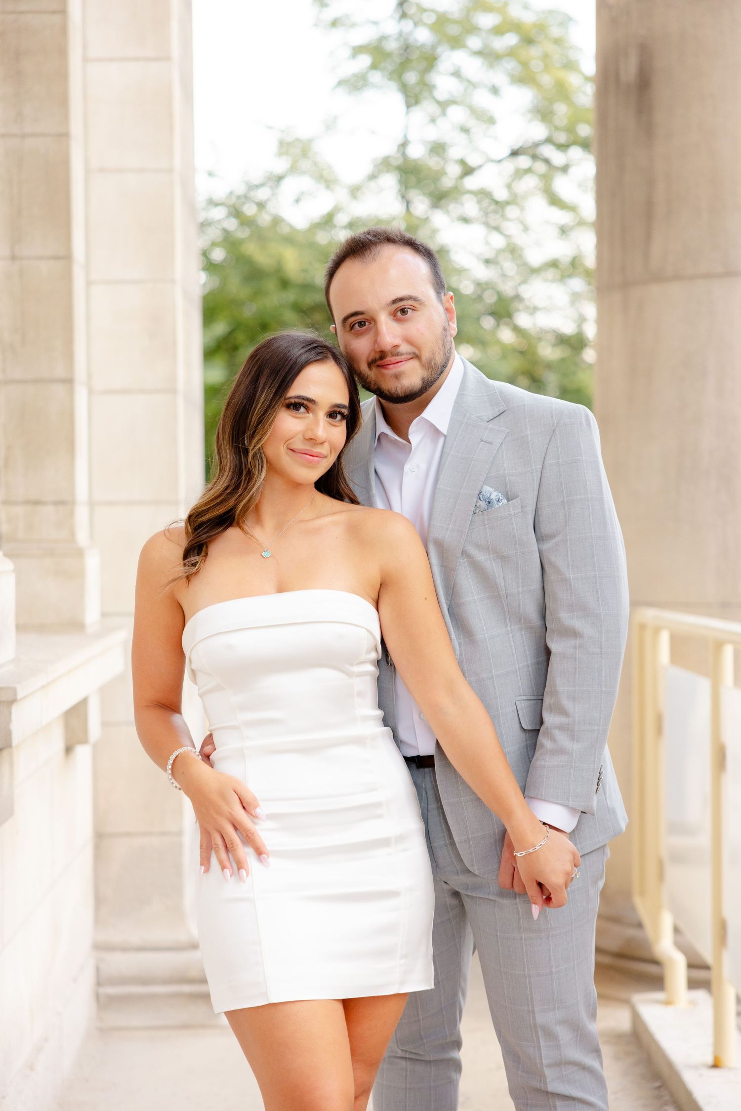 A romantic couple poses together on a covered terrace, with her in a white dress and him in a light gray suit.