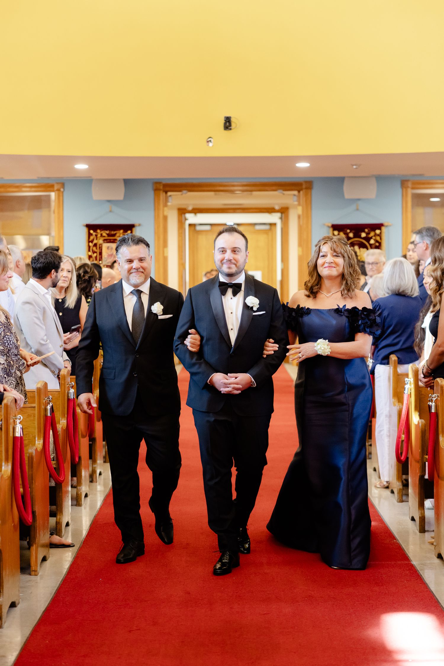 Wedding processional down red carpeted church aisle with attendants in formal navy blue attire.