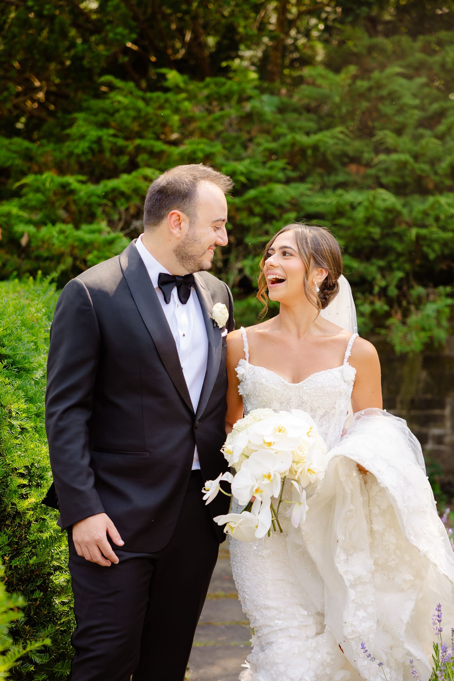 Happy newlywed couple laughing together outdoors with white orchid bouquet and formal wedding attire.