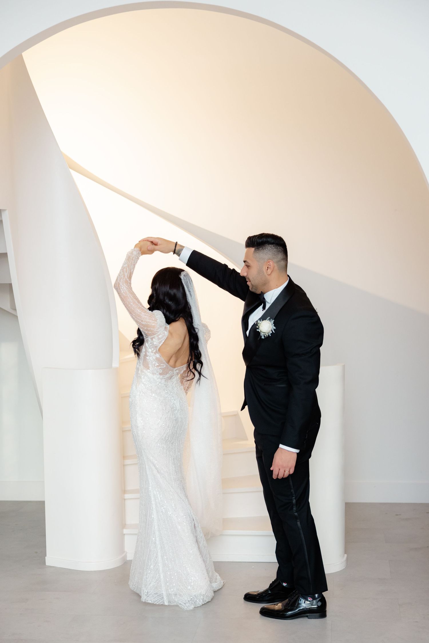 Newlyweds dance together on modern white staircase under curved archway.