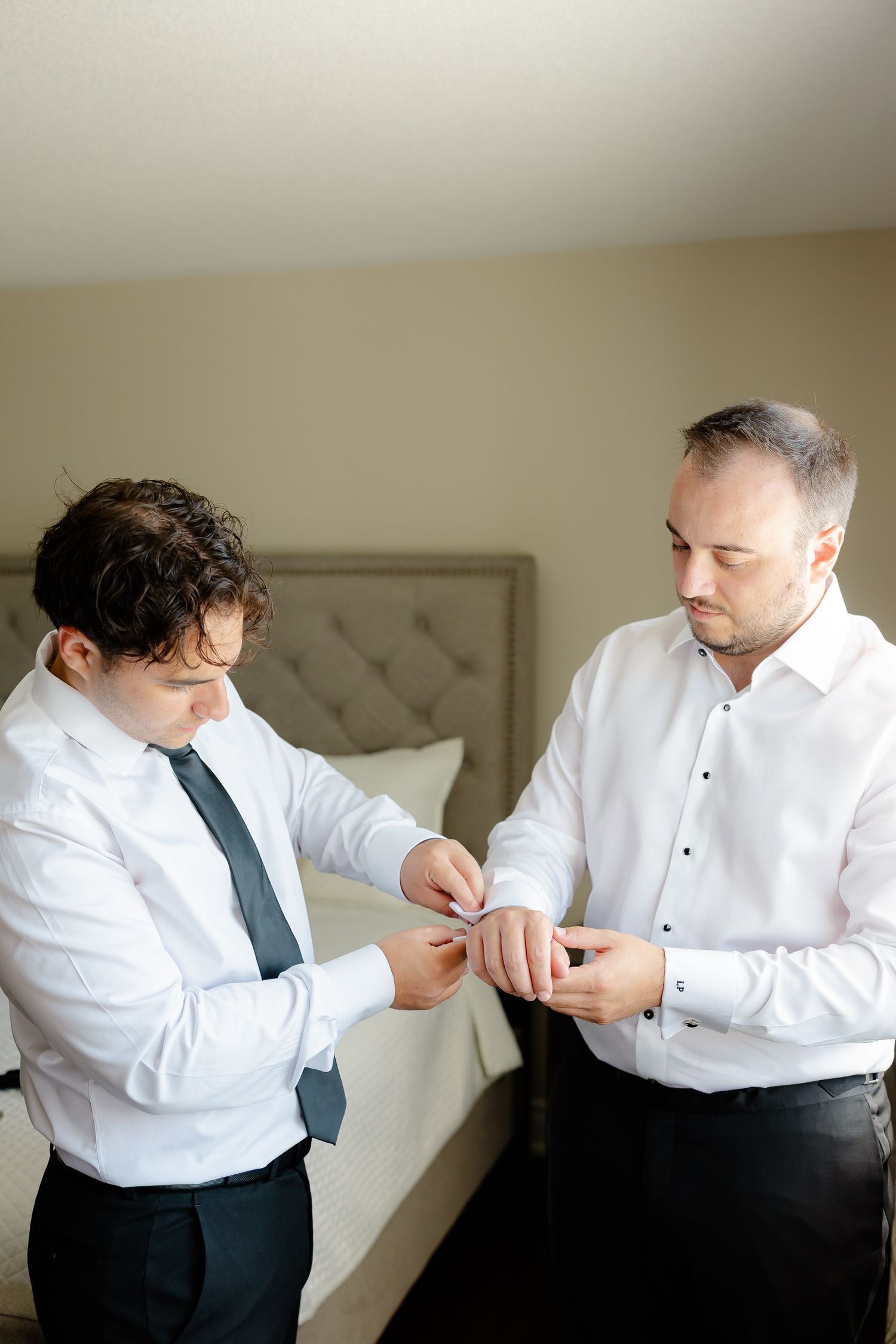 Two groomsmen in white shirts and black pants assist each other with formal wear preparations.