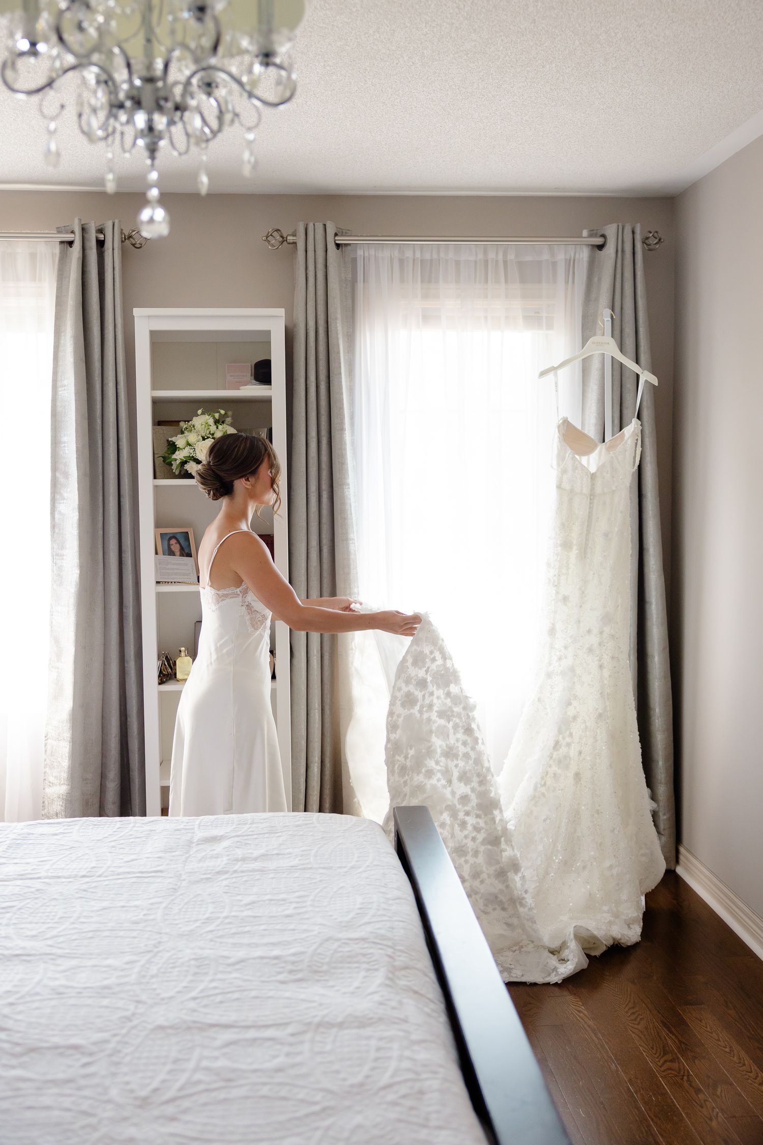 Two wedding dresses in an elegant bedroom with chandelier and gray curtains, one being worn and one hanging on display.