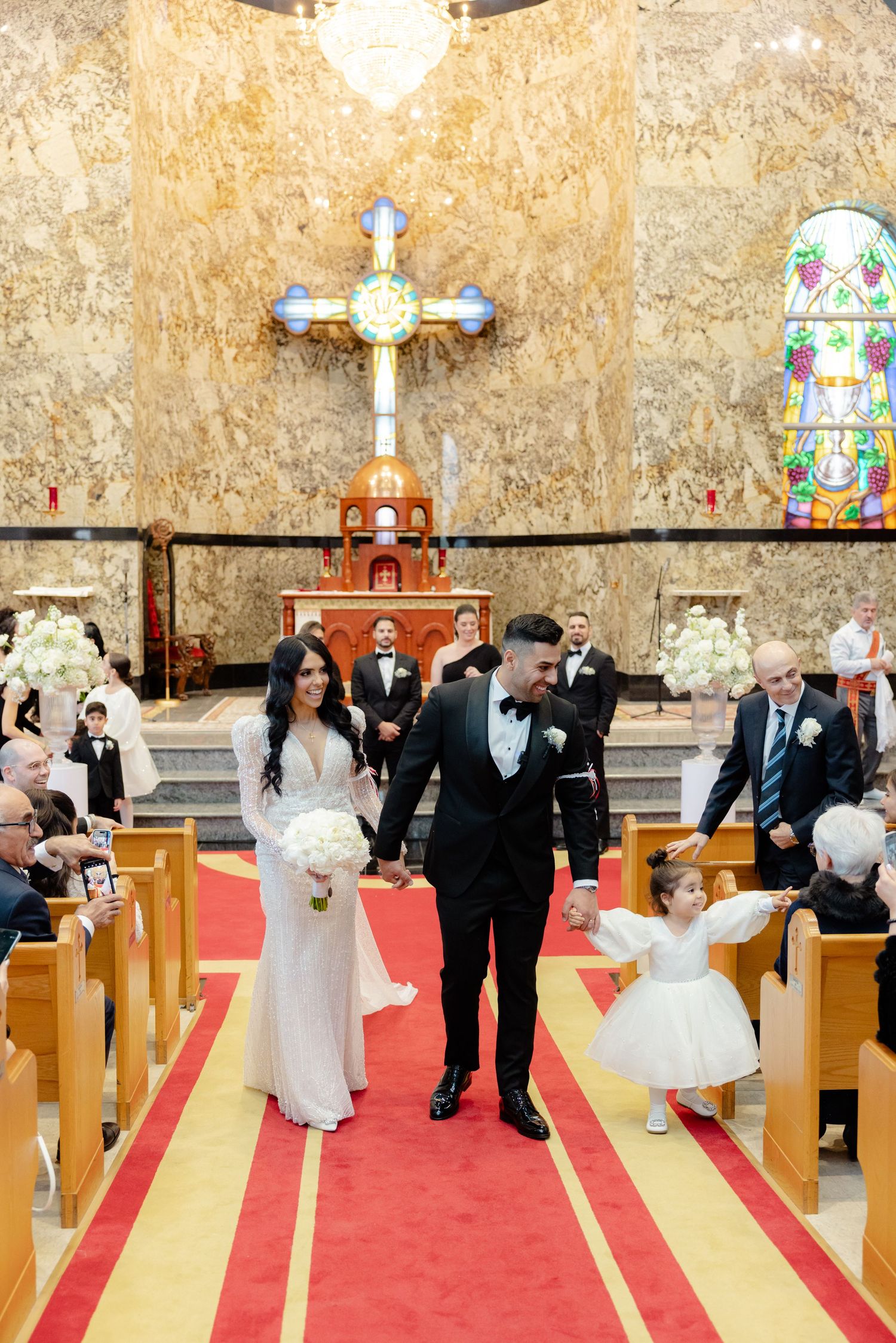 Wedding processional walks down red and gold carpeted church aisle beneath illuminated cross.