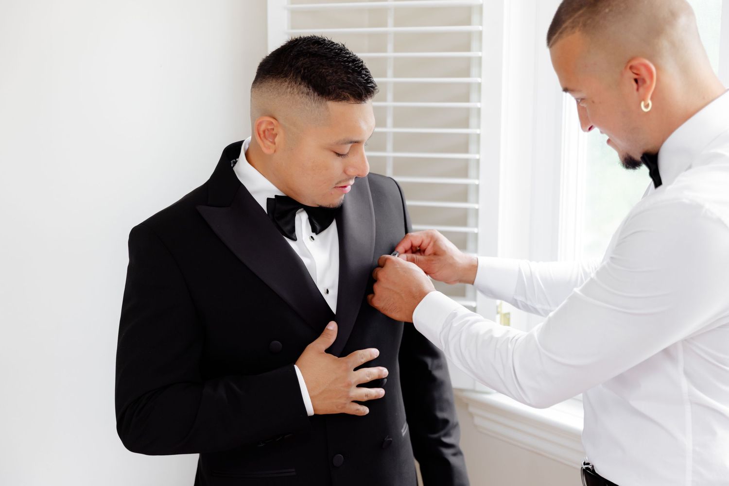 A groom adjusts his black suit jacket in a well-lit room while preparing for his wedding day.