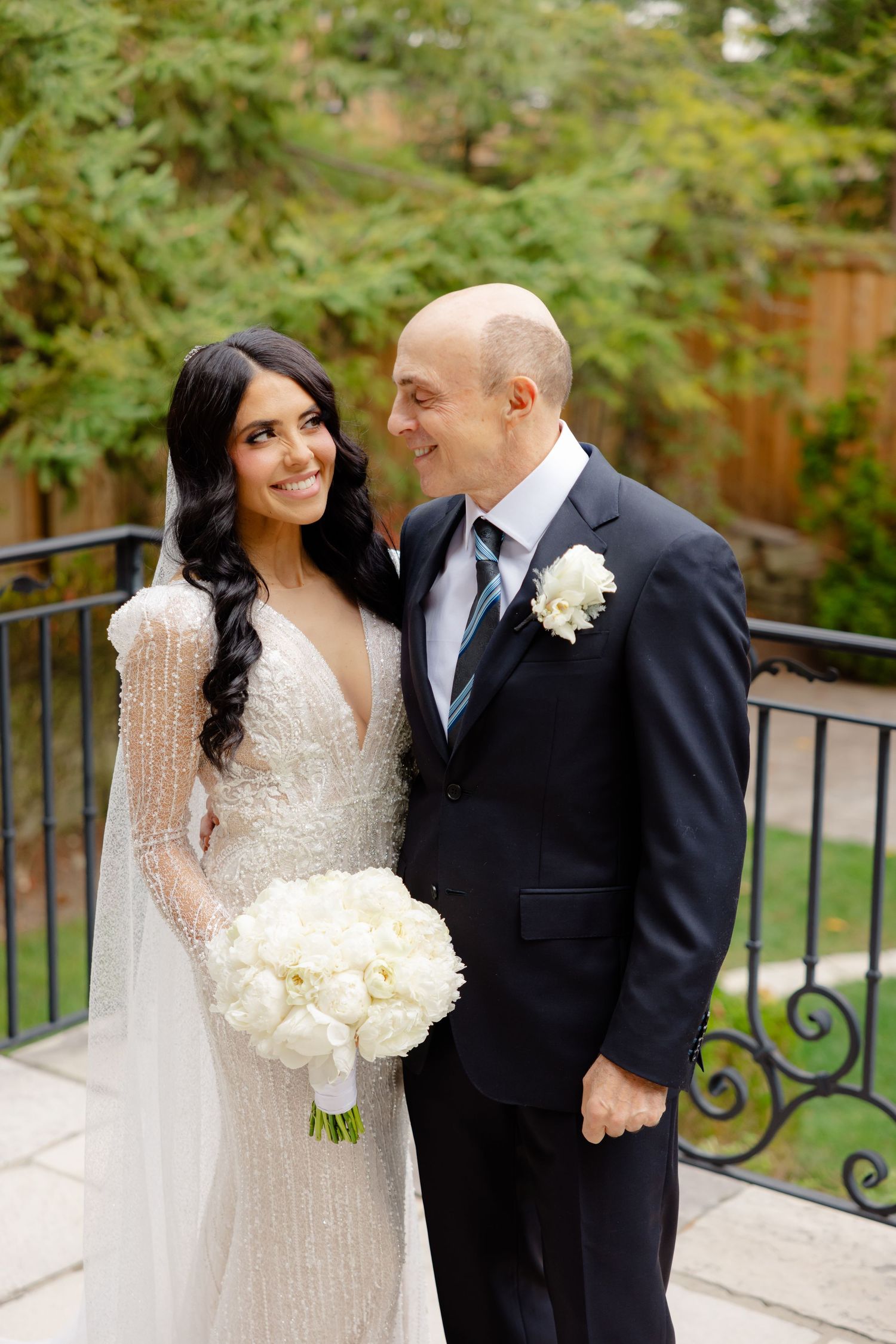 Elegant wedding couple poses outdoors, with bride in beaded gown holding white bouquet and groom in classic dark suit.