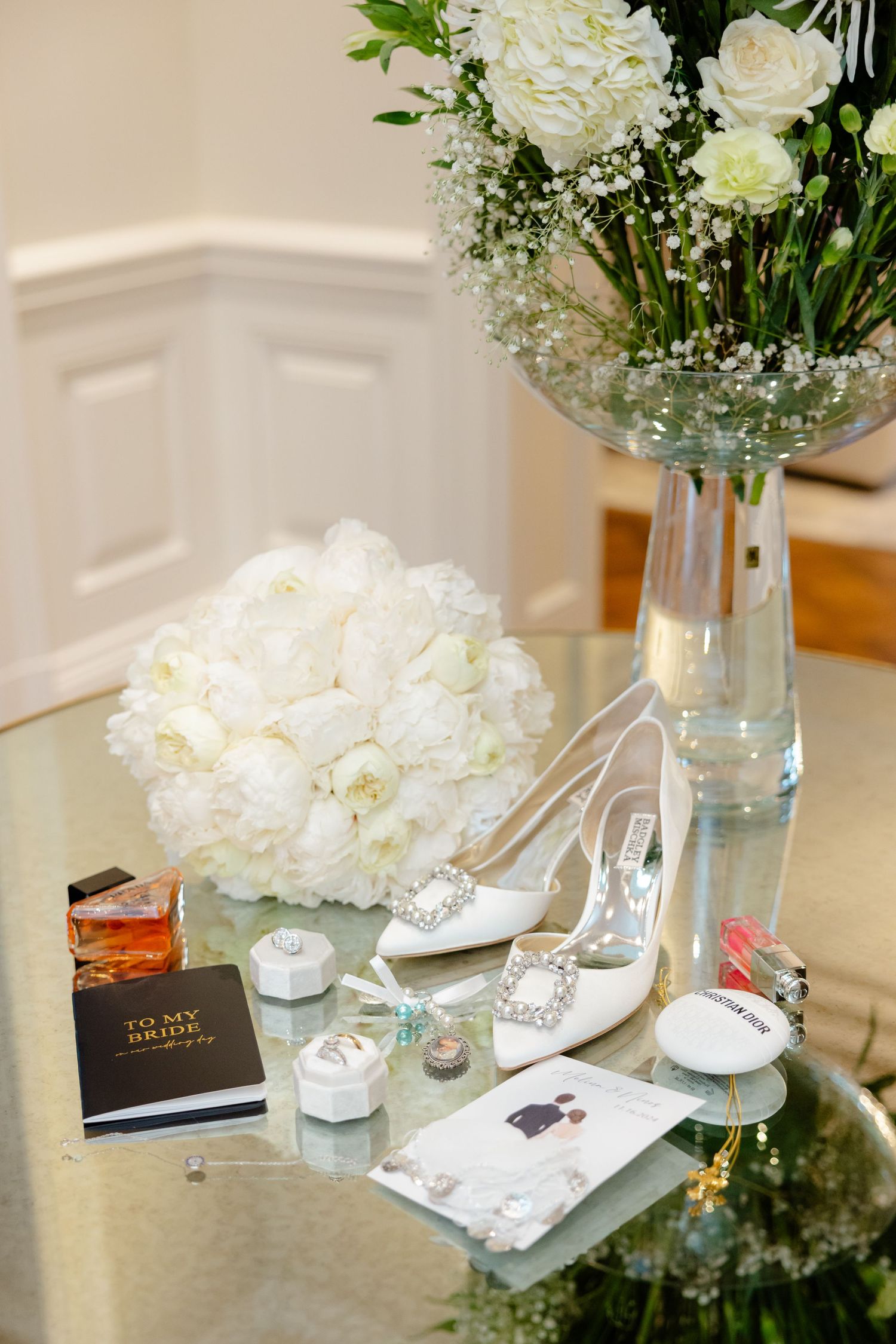 Wedding detail flatlay with white flowers, bridal shoes, and stationery on reflective surface.
