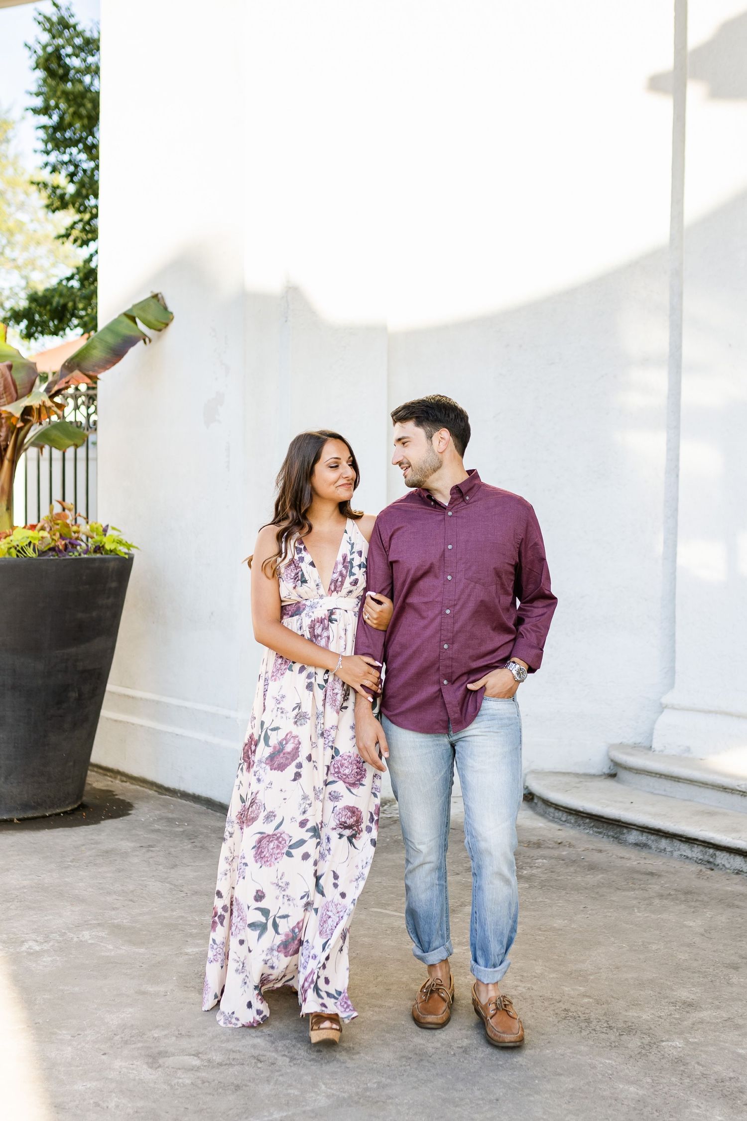 A couple stands together against a white wall wearing coordinated purple and floral outfits.