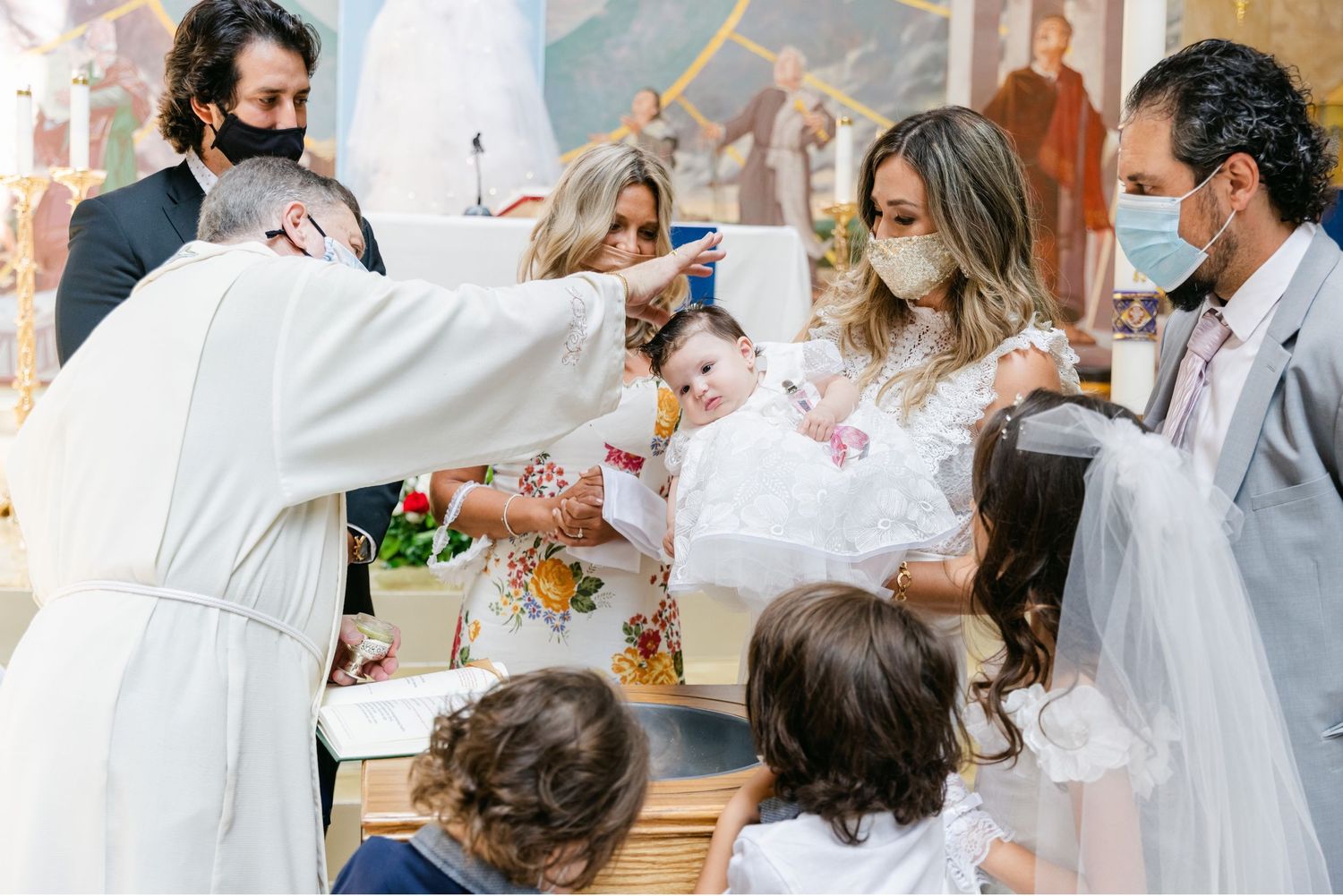 A priest performs baptismal rites while family members watch in a church setting.