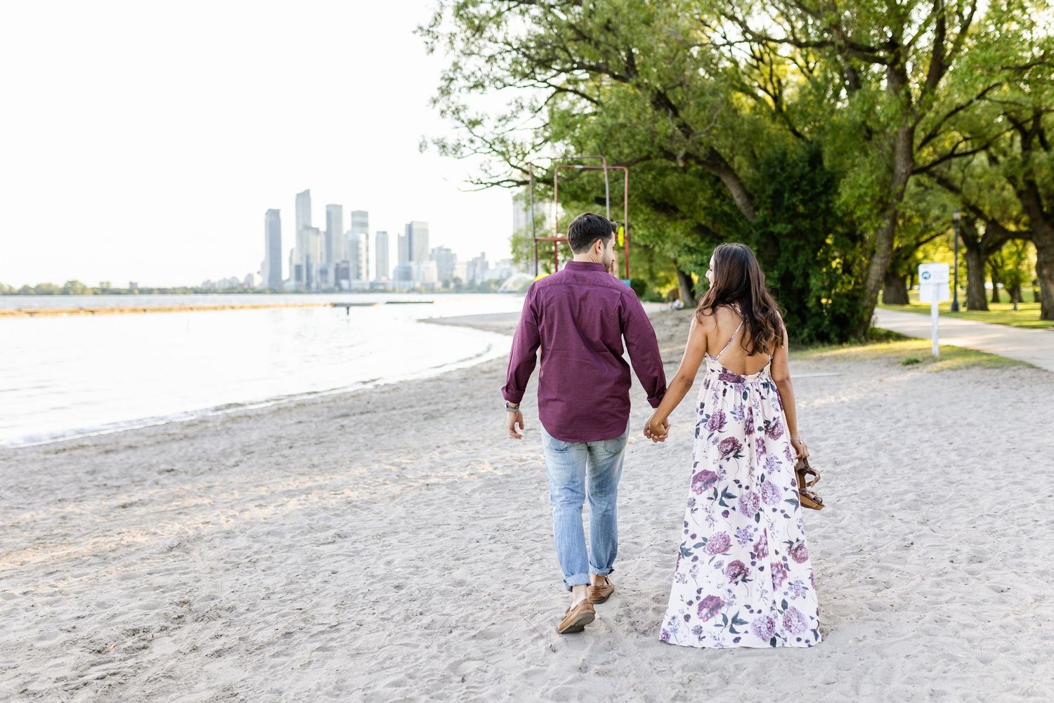 Couple walking hand in hand on beach with city skyline in background.