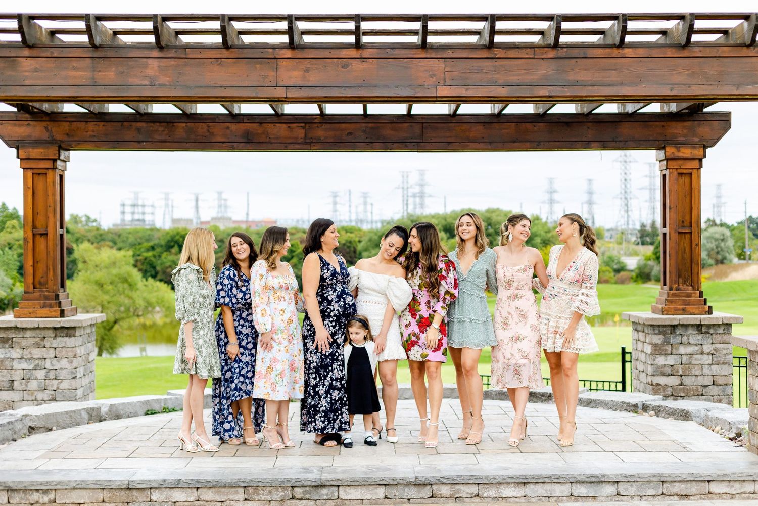 Ladies in summer dresses pose together on a stone patio under a wooden pergola overlooking scenic landscape.