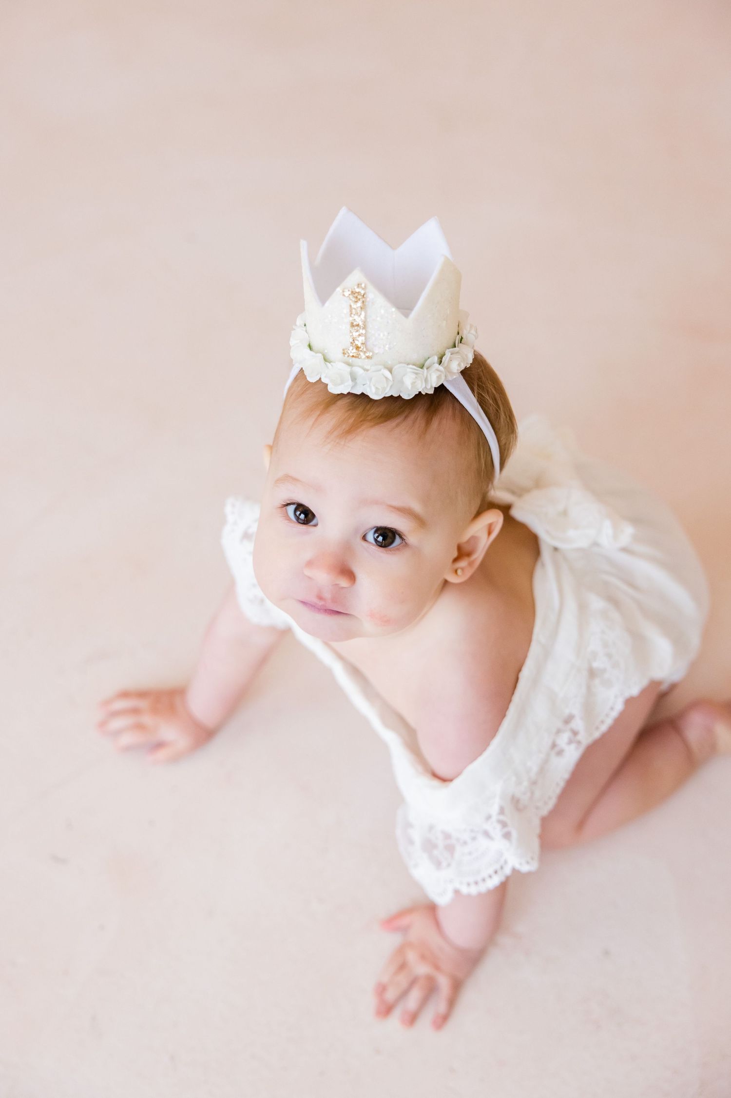 High angle portrait with delicate white crown and lace dress against pink background.
