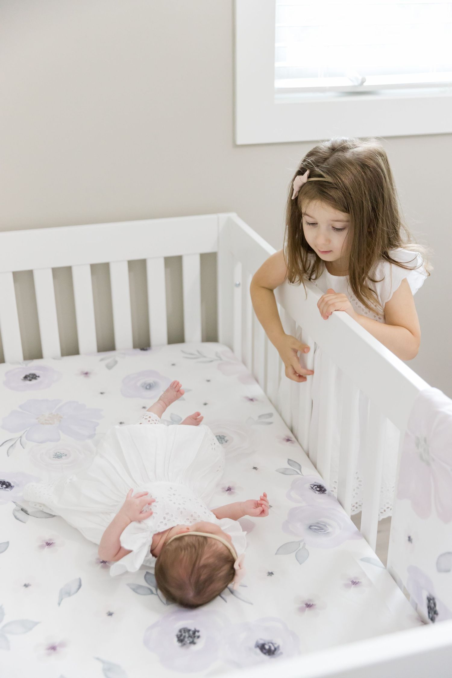 Sibling watches over baby lying in white crib with floral bedding.