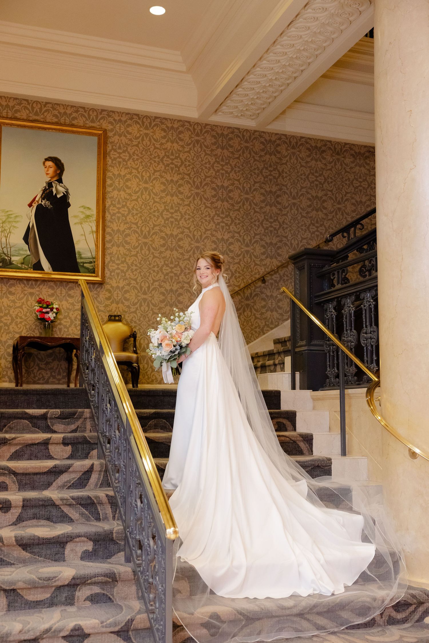 A bride in a cathedral-length veil descends an ornate staircase with gold railings and patterned carpet.