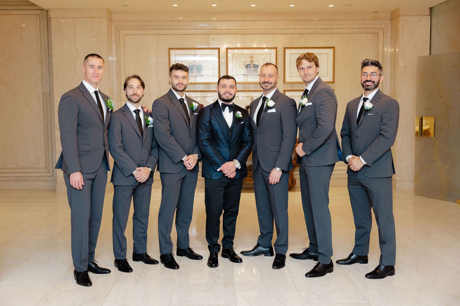 Groomsmen in matching dark suits stand in formation against a neutral ballroom backdrop.
