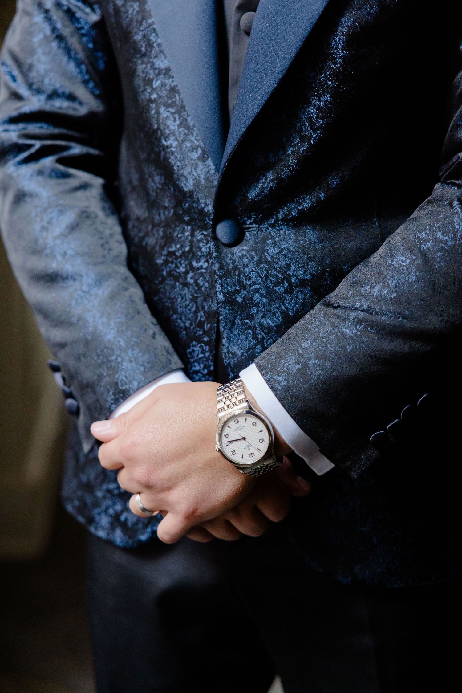 Close-up view of a silver wristwatch with a metallic band worn with a dark textured formal suit jacket.