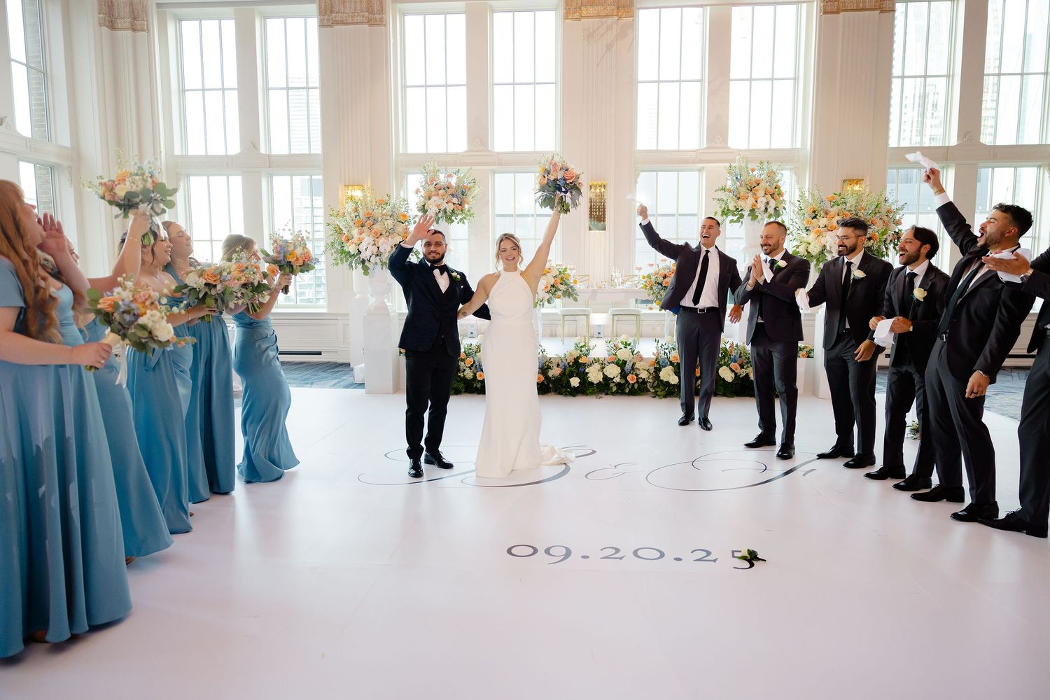 Wedding party lined up in formal attire against white walls with large windows.