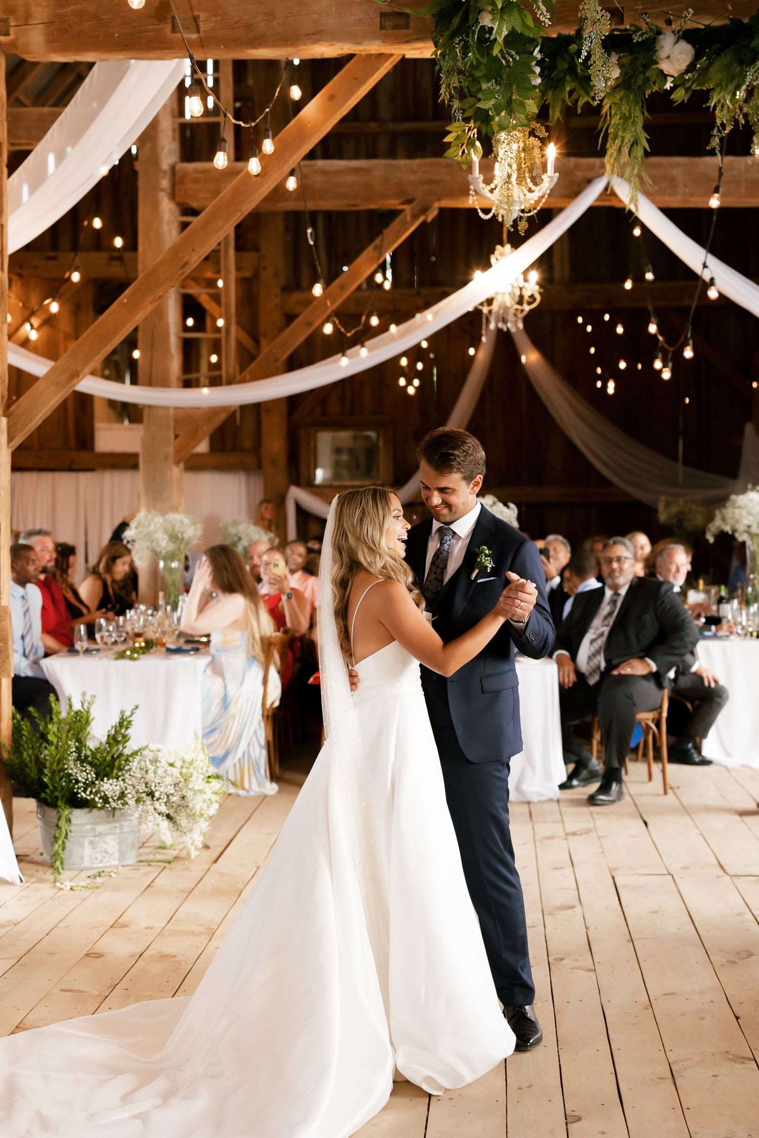 Romantic dance in rustic barn venue decorated with elegant white draping and chandeliers.