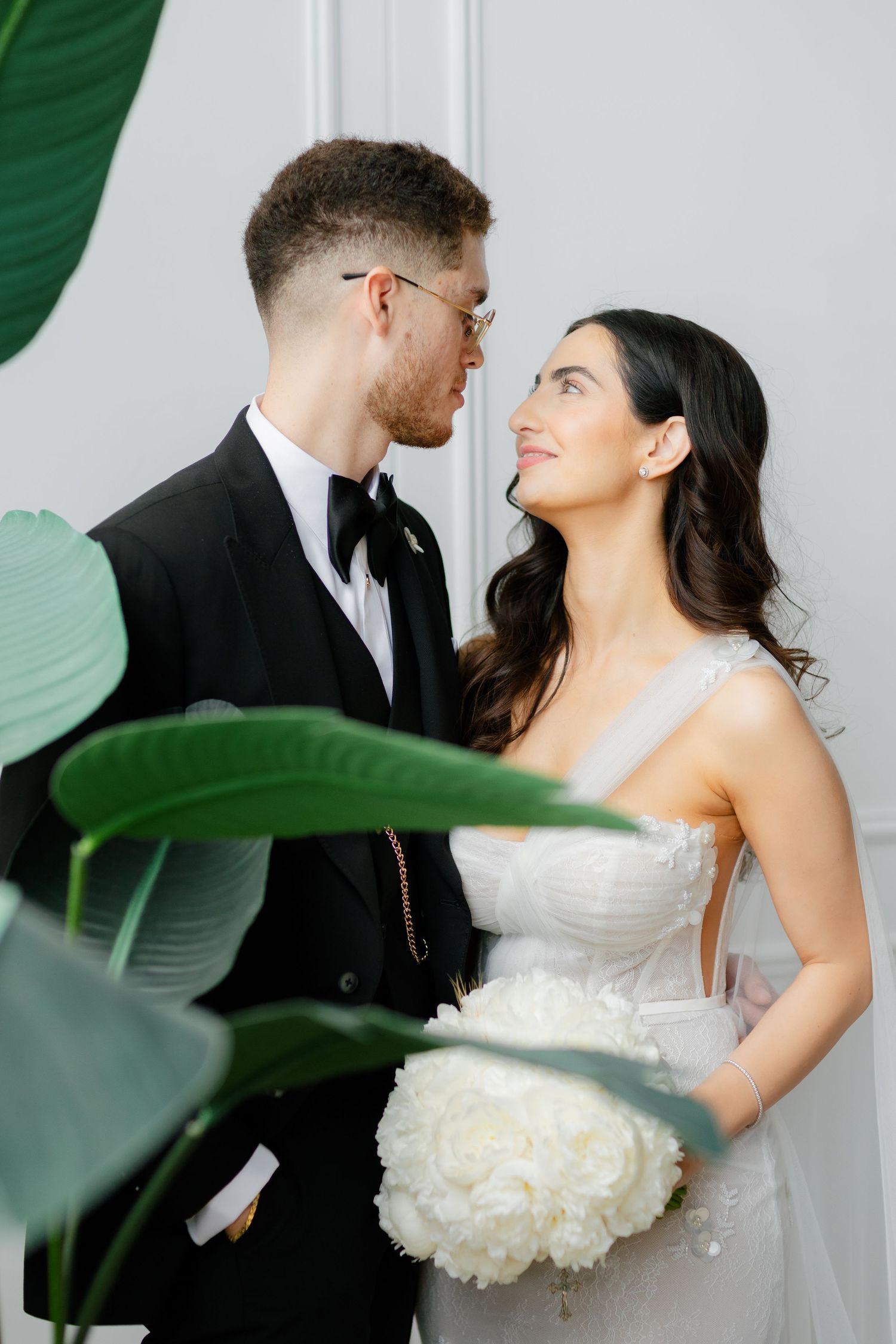 Elegant wedding portrait with couple and tropical leaves against white wall.