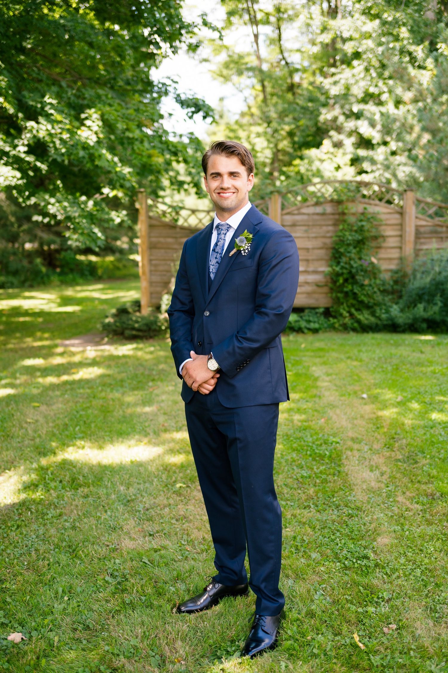 Formal portrait in navy three-piece suit with boutonniere in garden setting.