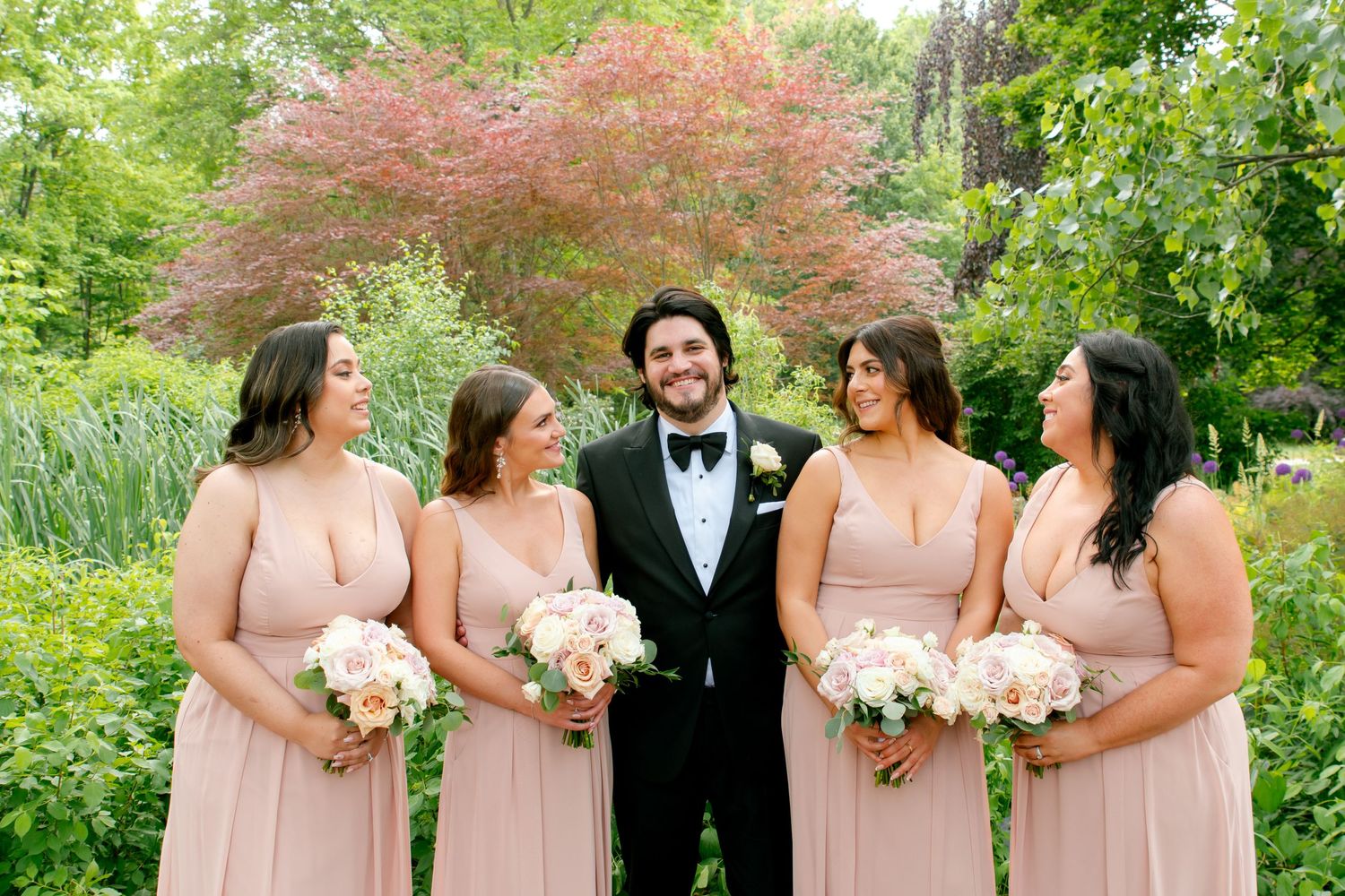 Wedding party photo featuring bridesmaids in pink dresses with a groom against a flowering garden backdrop.