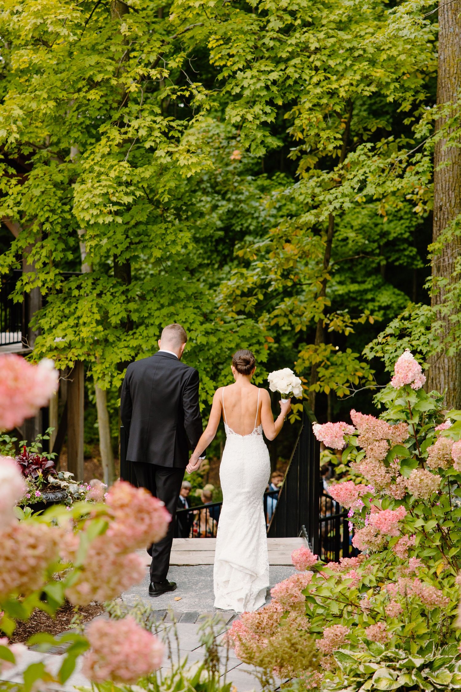 Wedding party walks along garden path surrounded by blooming hydrangeas and green foliage.