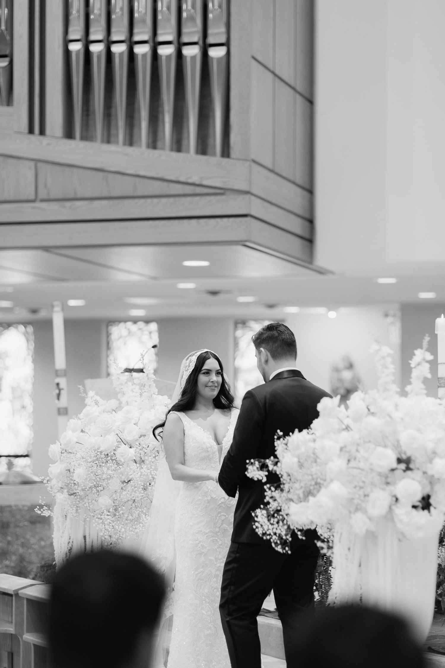 Black and white photo of wedding couple with white floral arrangements in church.