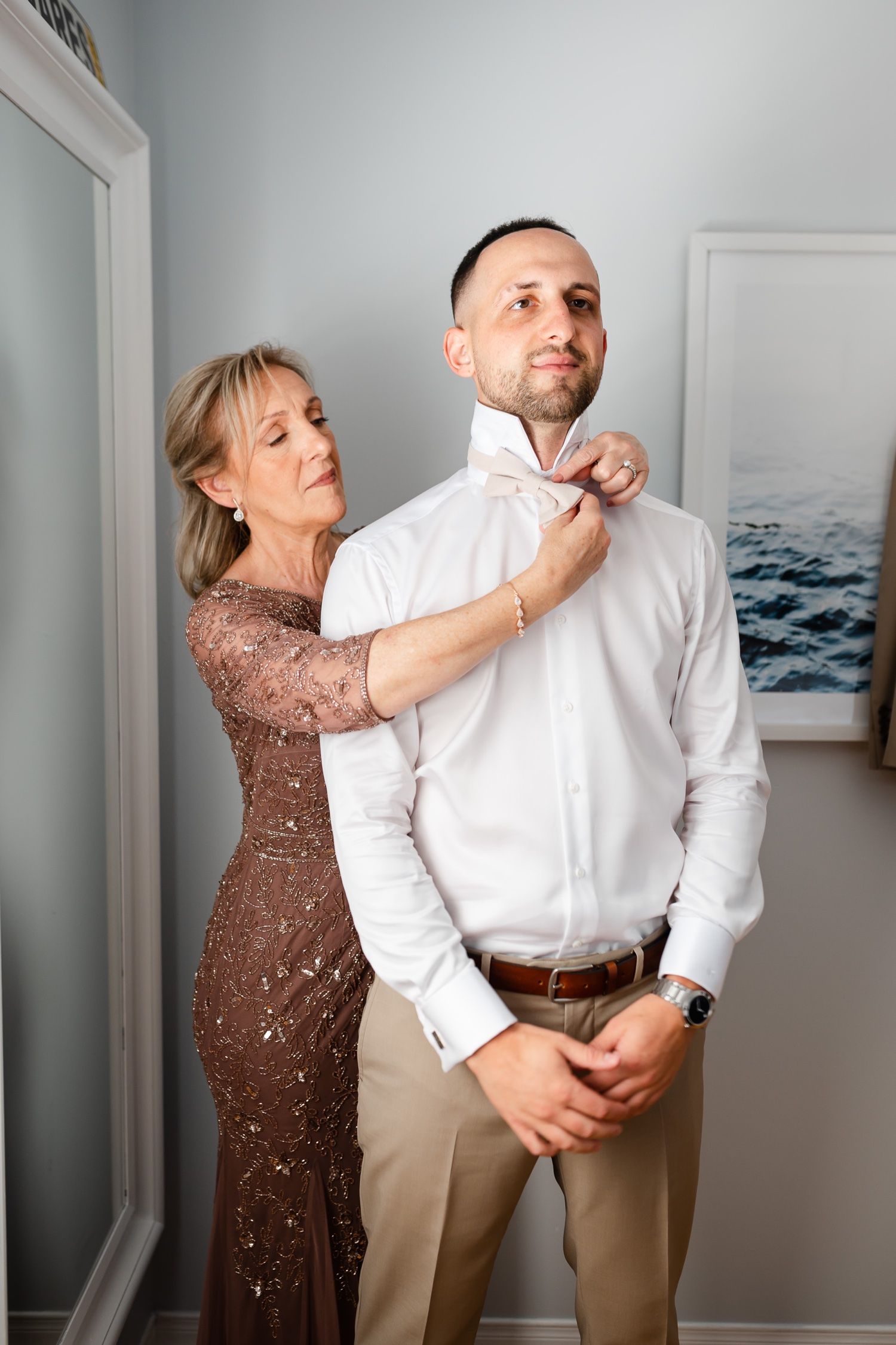 A person in a sparkly brown dress helps adjust another's white collar during wedding preparations.