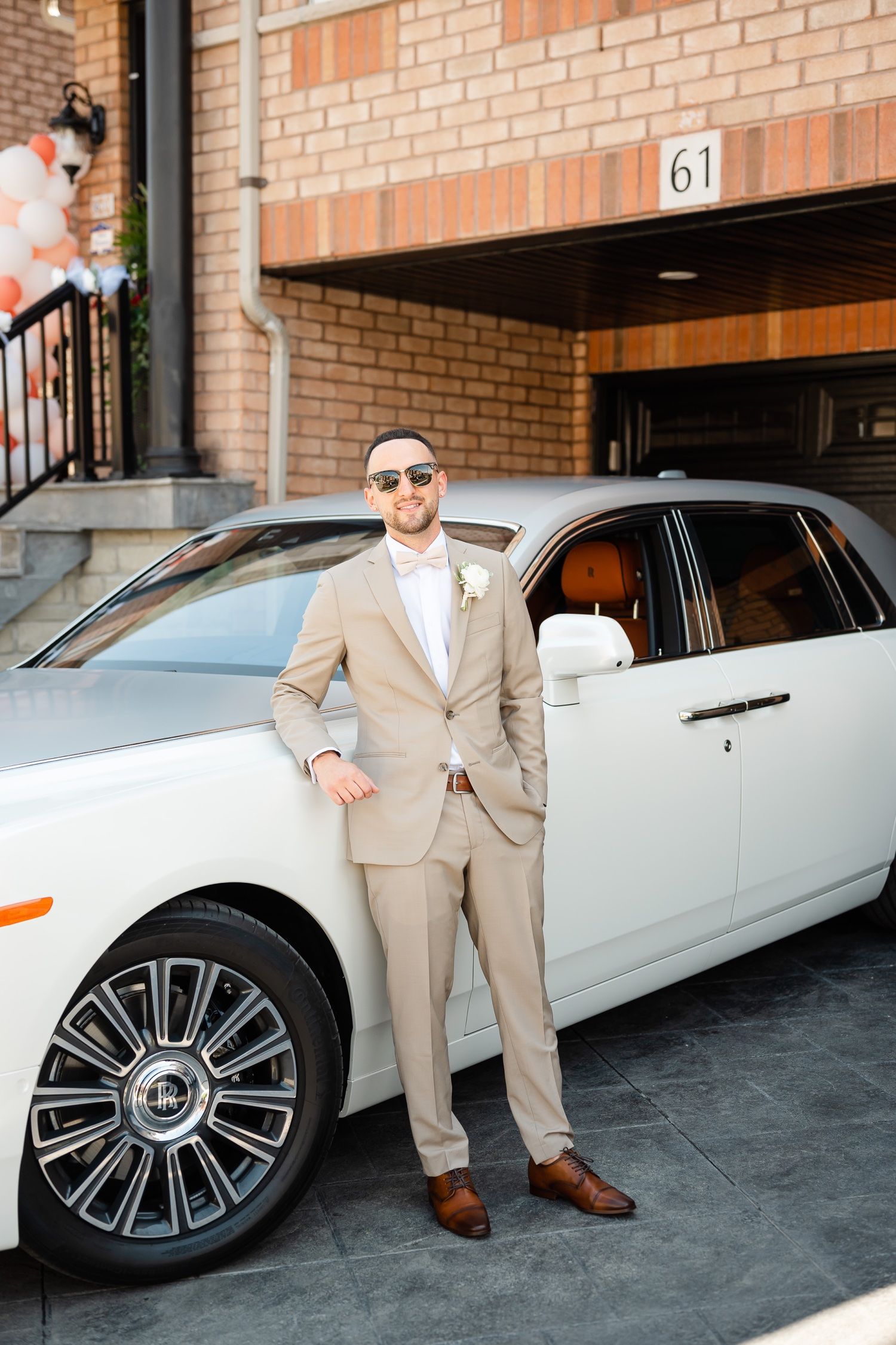 Stylish groom wearing sunglasses stands beside white luxury vehicle at brick building entrance.