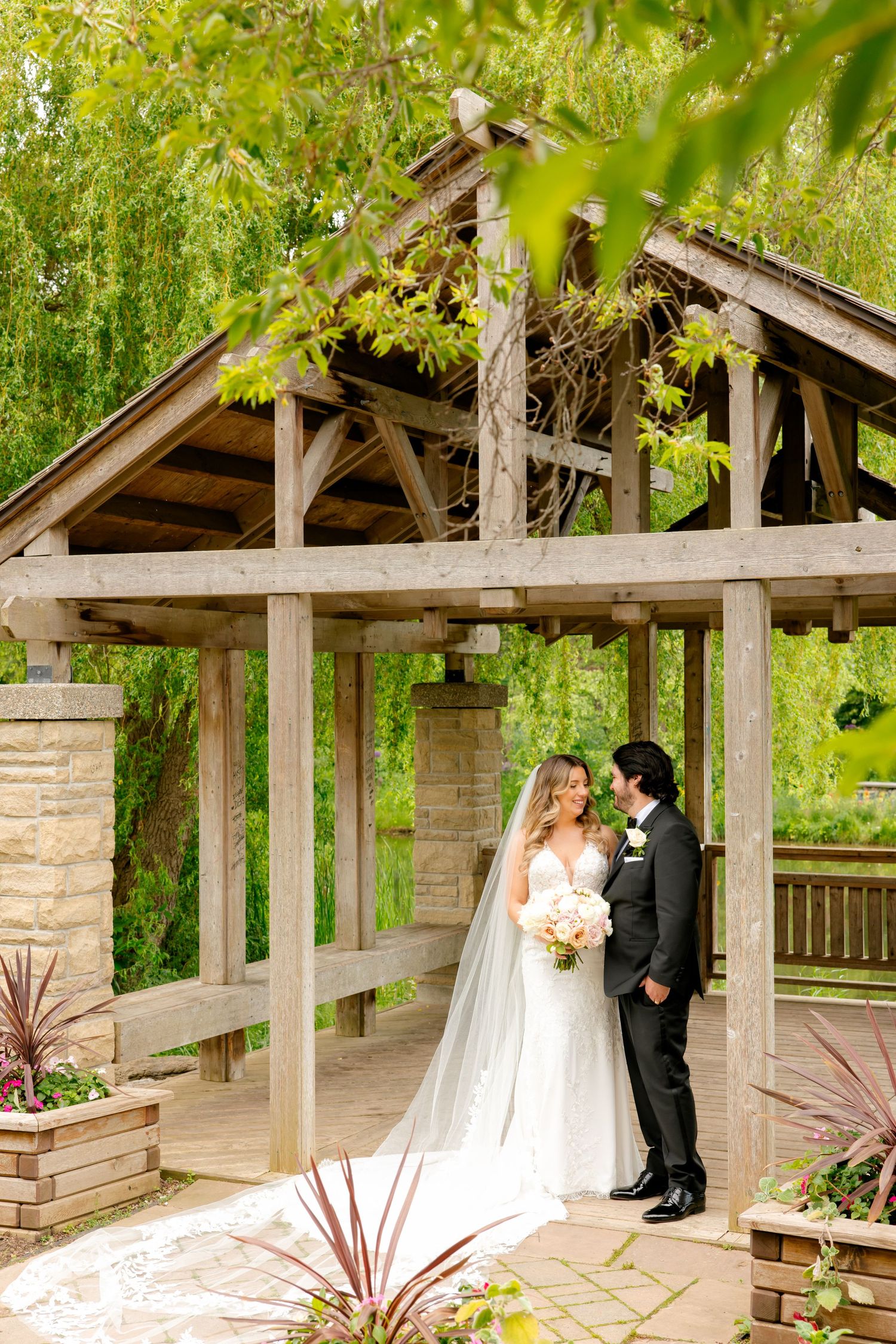 Romantic wedding portrait of couple embracing under rustic wooden gazebo in garden setting.