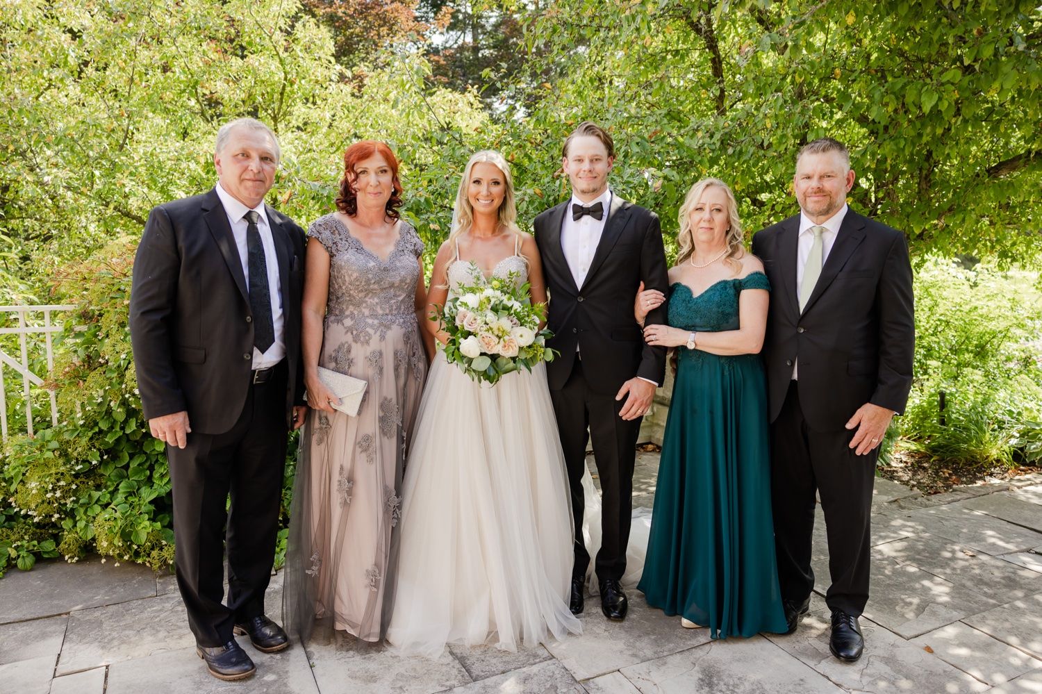 Formal family portrait of wedding party on a stone patio surrounded by greenery.