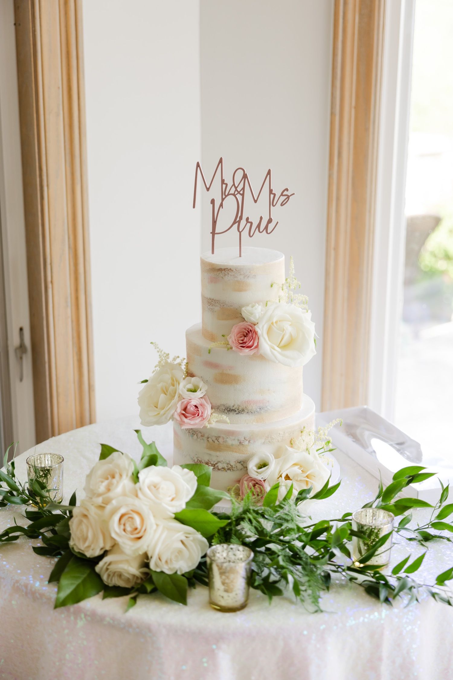 Three-tiered naked wedding cake adorned with fresh roses and greenery on white tablecloth.