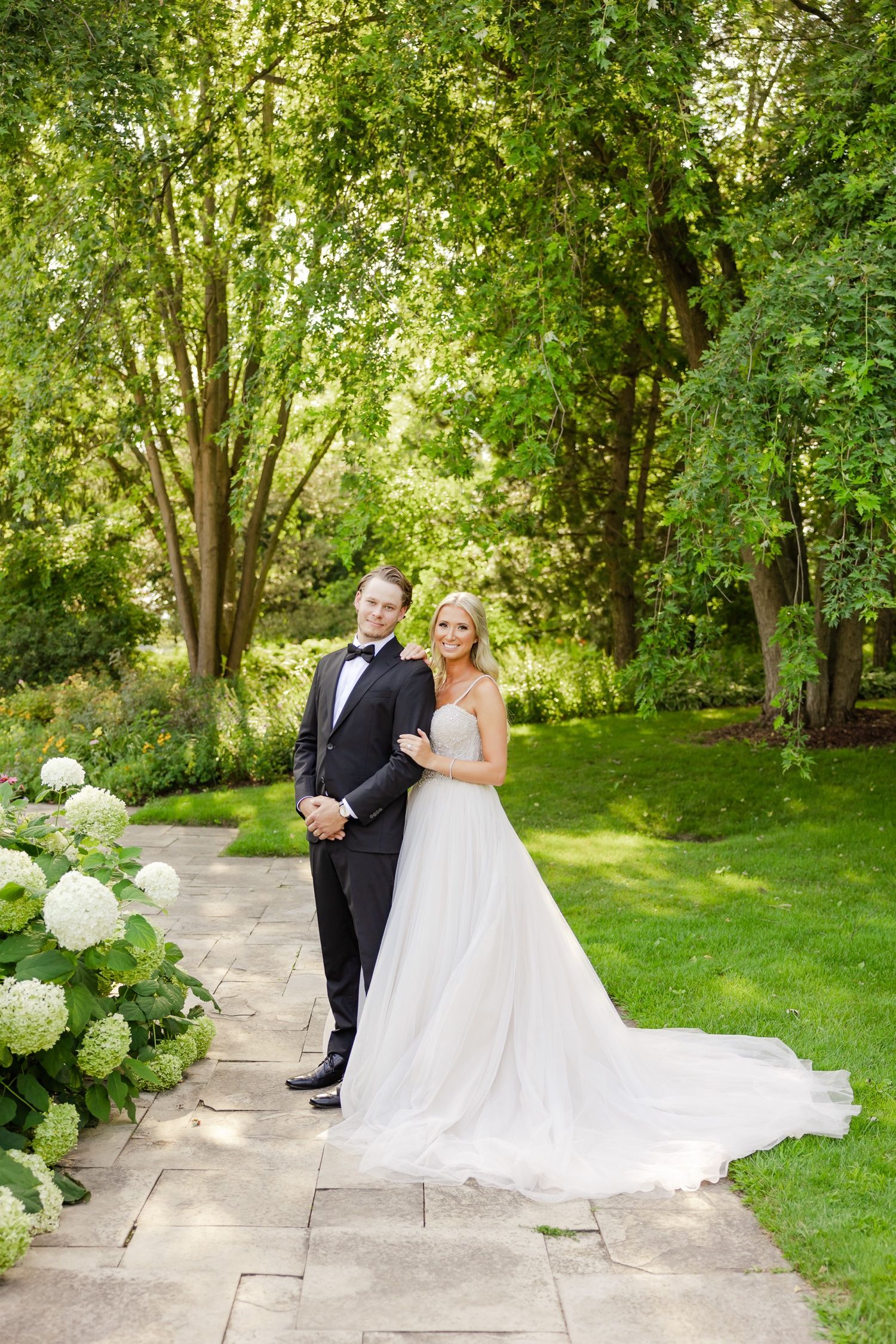 Wedding couple poses on stone pathway with flowing bridal gown and garden backdrop.