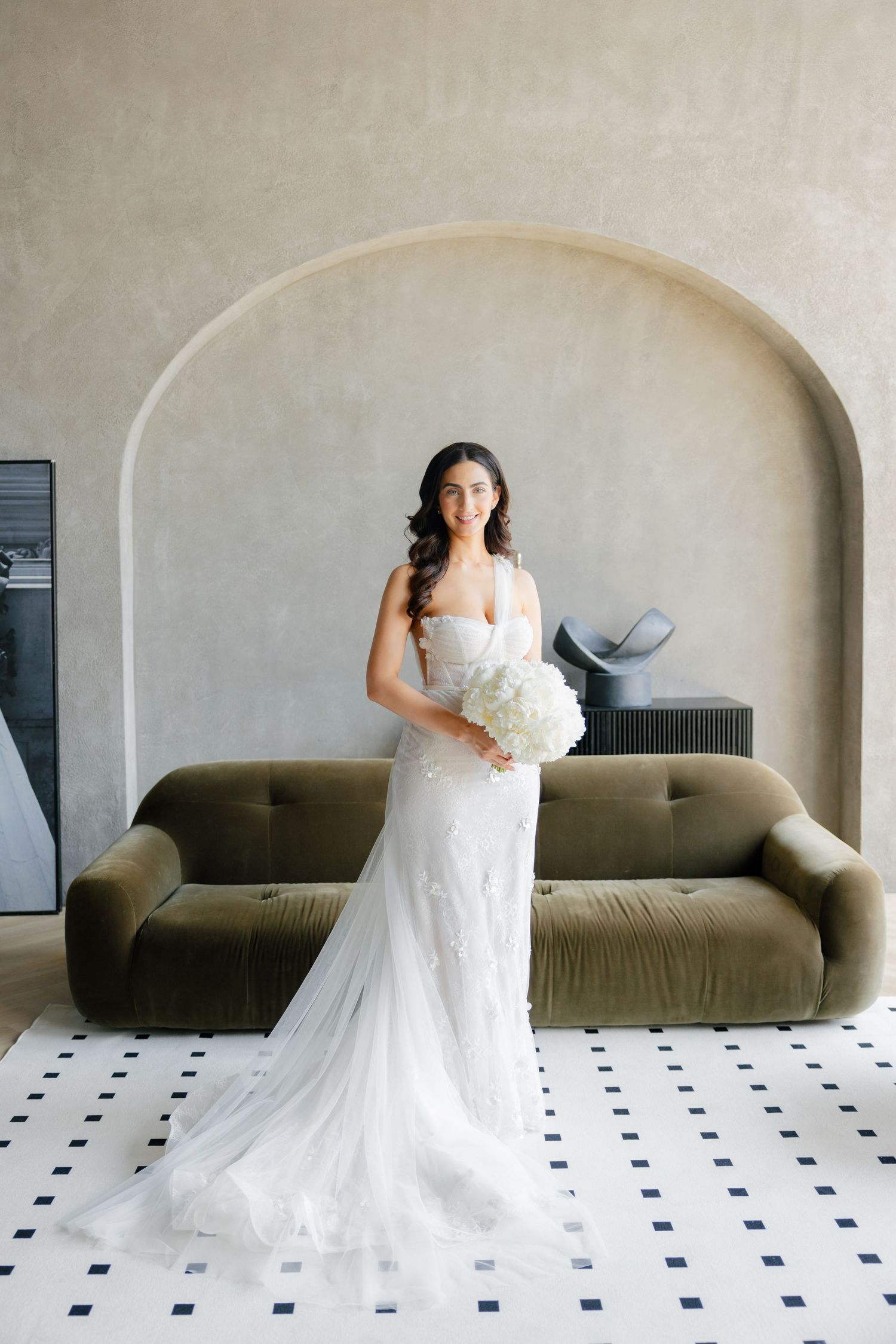 Bride in fitted lace gown holds white bouquet while standing in front of arched wall and olive sofa.