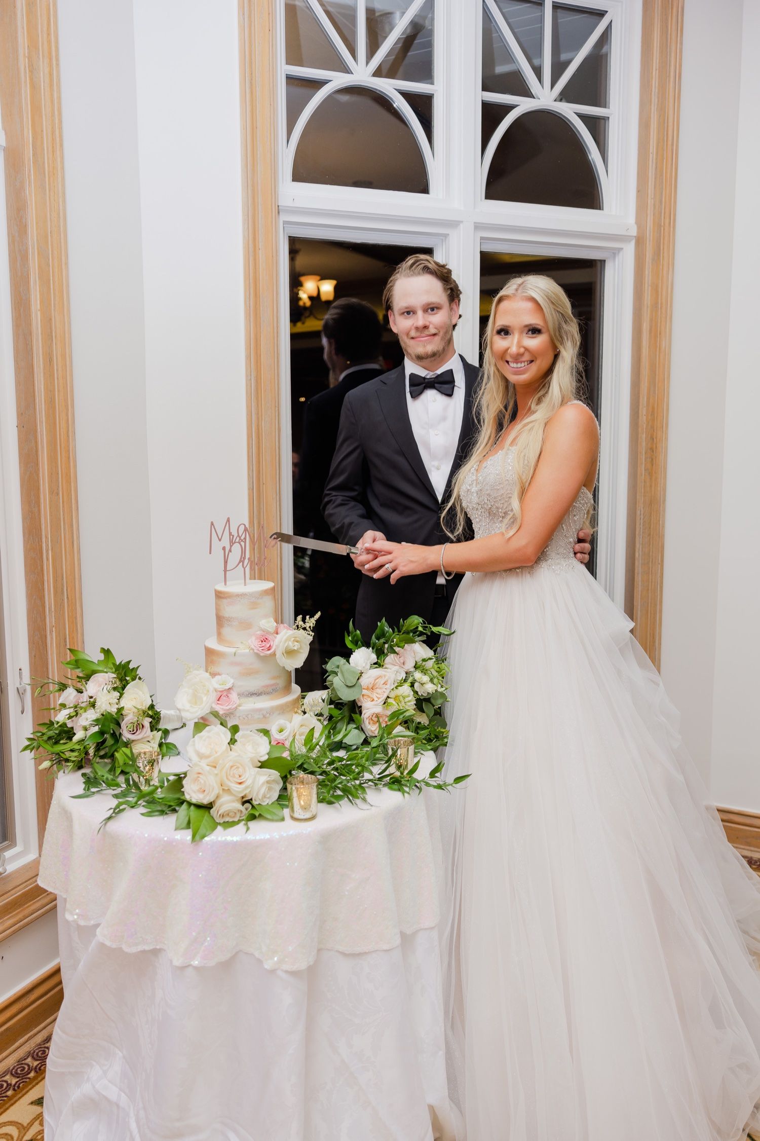 Wedding couple stands beside an elegantly decorated cake table with white floral arrangements.
