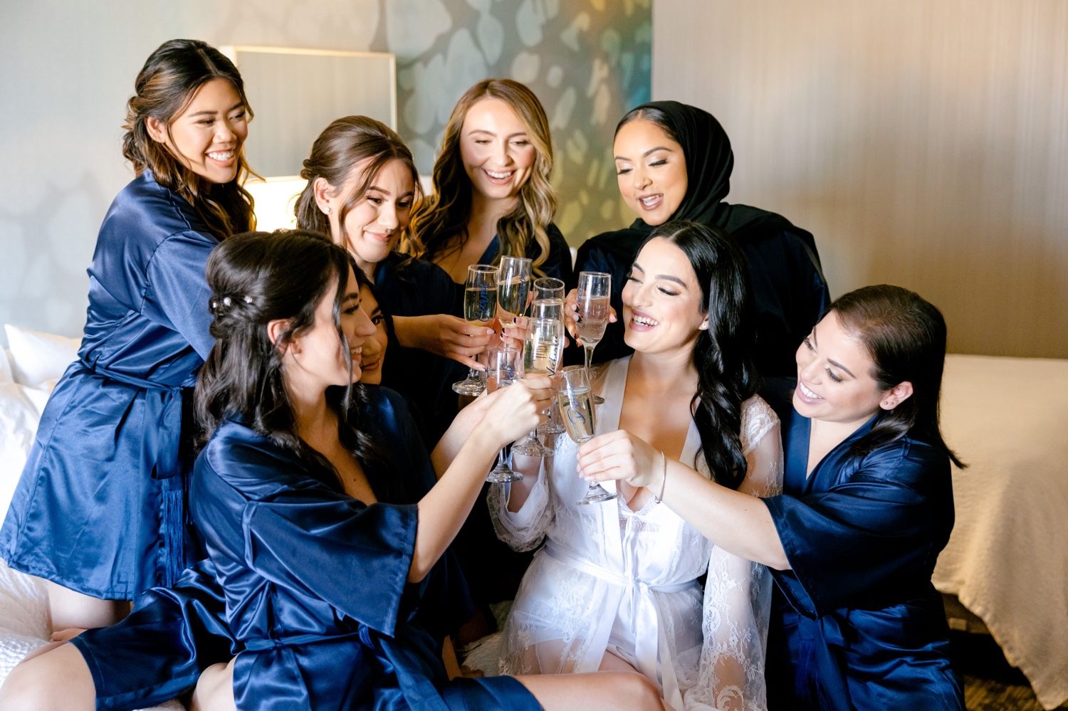 Bridesmaids in navy robes toast champagne glasses with the bride in white during pre-wedding preparations.