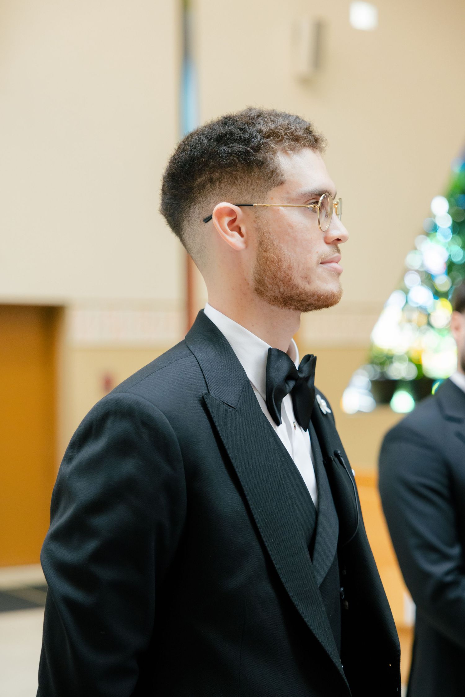 Groom in a classic black tuxedo and bow tie looks to the side while standing in front of a Christmas tree.