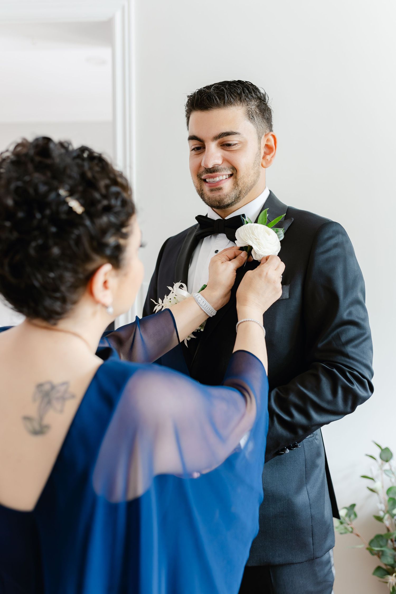A white boutonniere is pinned onto a black tuxedo jacket during wedding preparations.