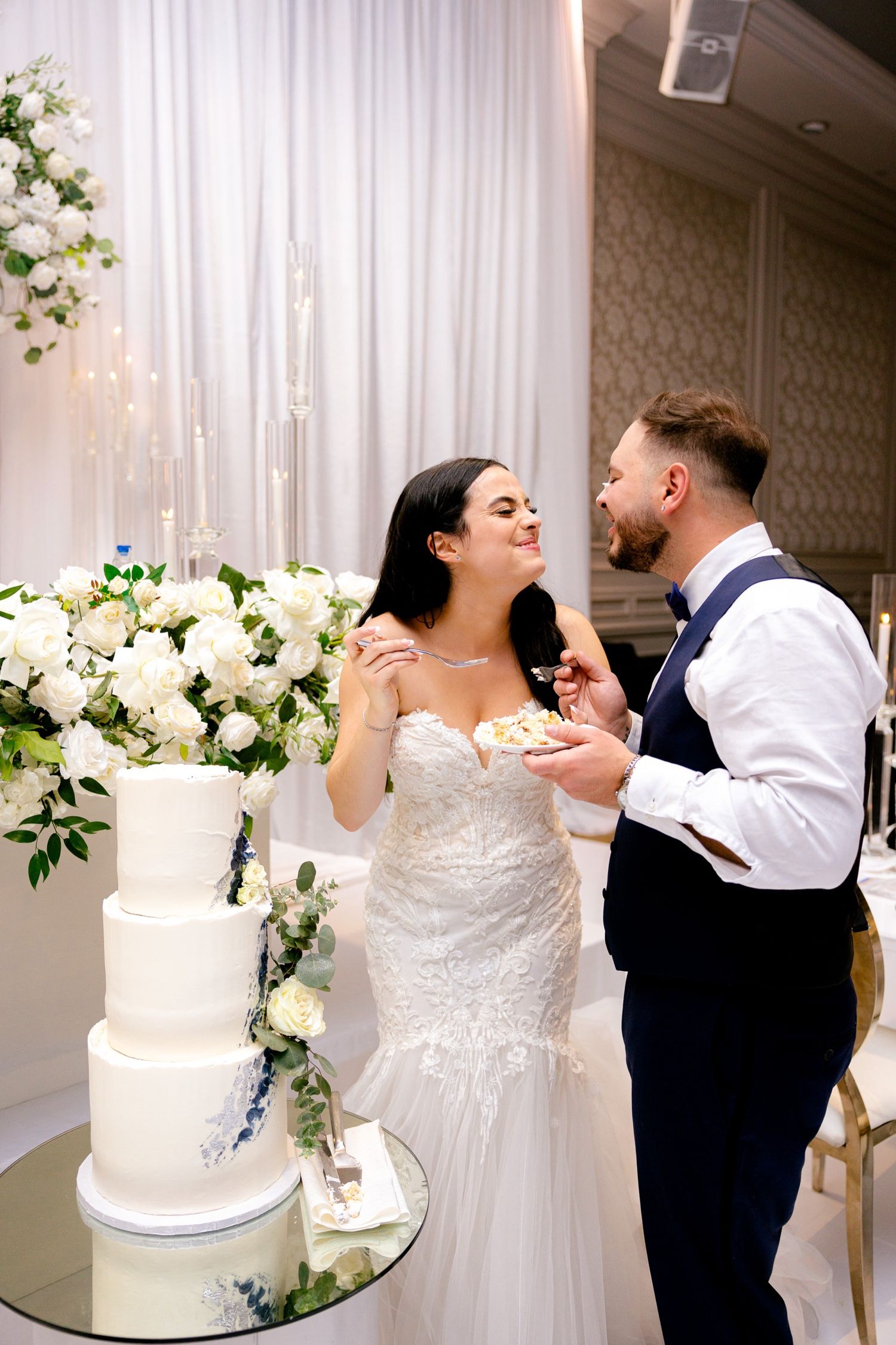 Newlyweds playfully feeding each other wedding cake beside elegant white tiered dessert.