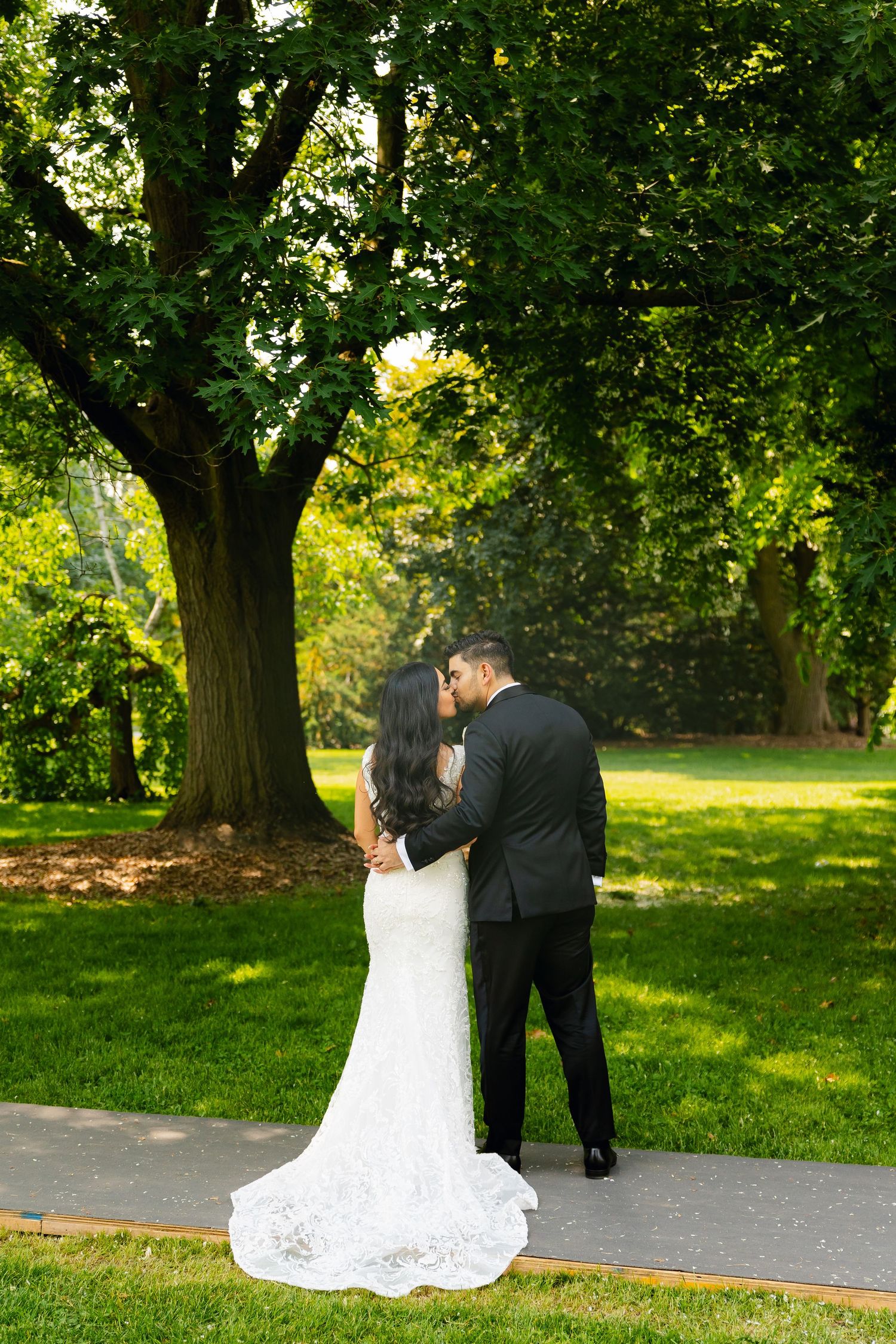 A couple in formal wedding attire share an intimate moment under a lush green tree.