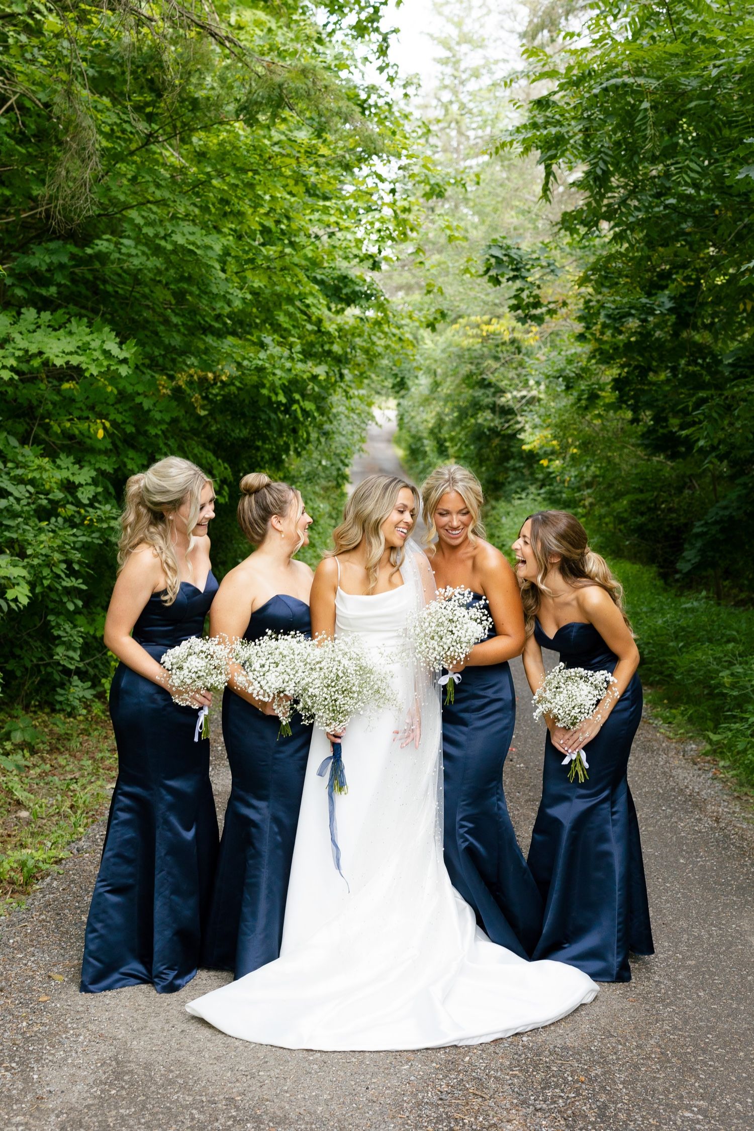 Bridal party sharing candid moment and laughter on wooded path in navy and white gowns.