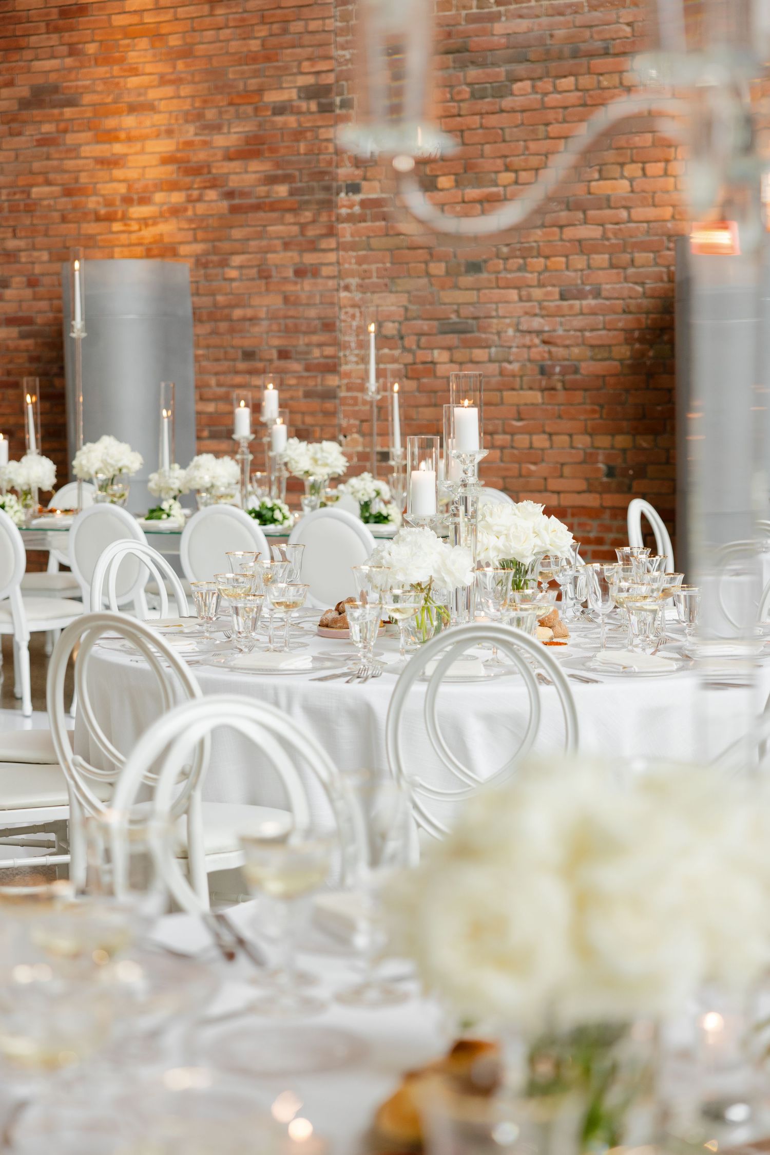 White and silver wedding table settings with floral centerpieces against exposed brick walls.