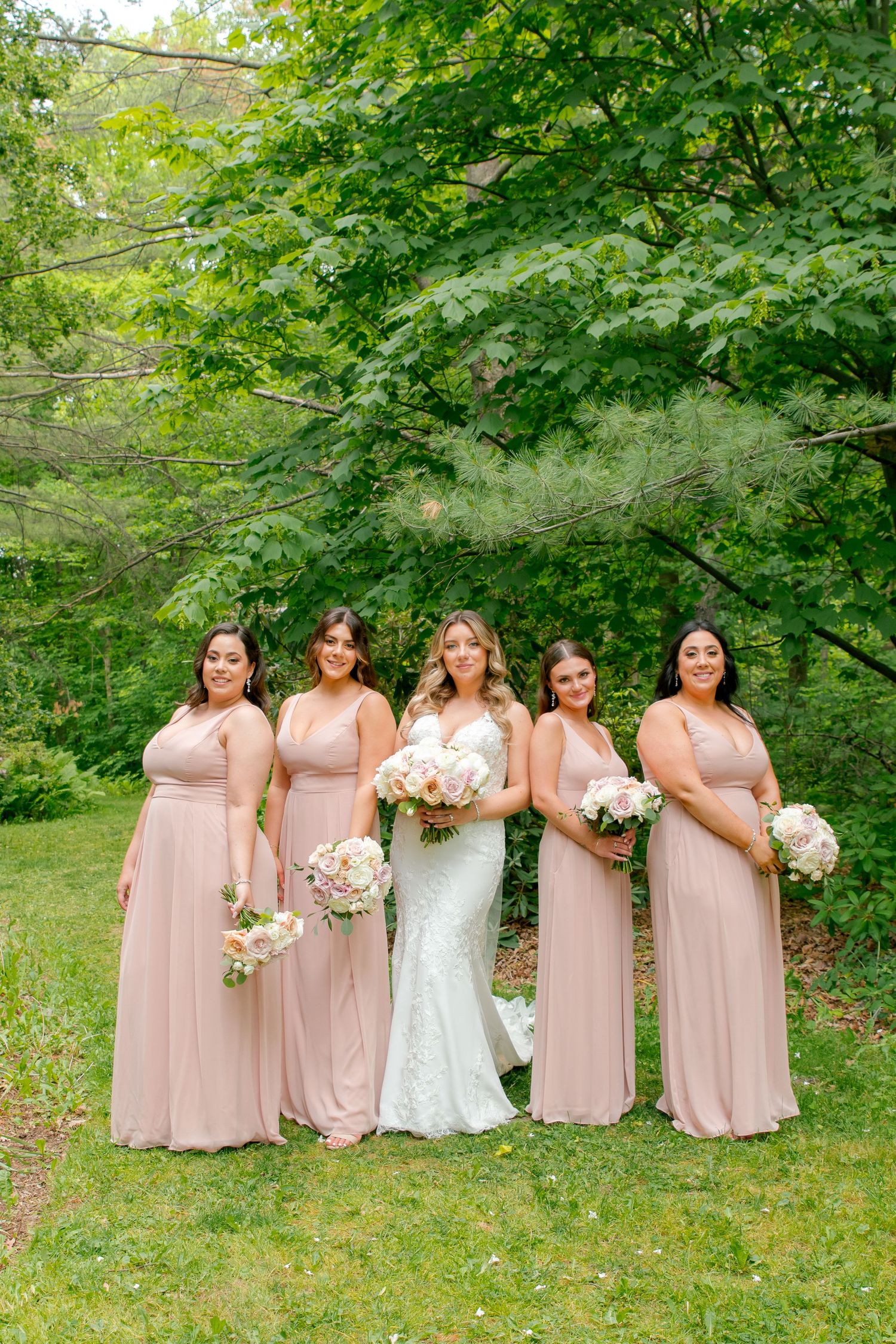 Bride in white gown stands with bridesmaids wearing matching blush pink dresses holding bouquets.