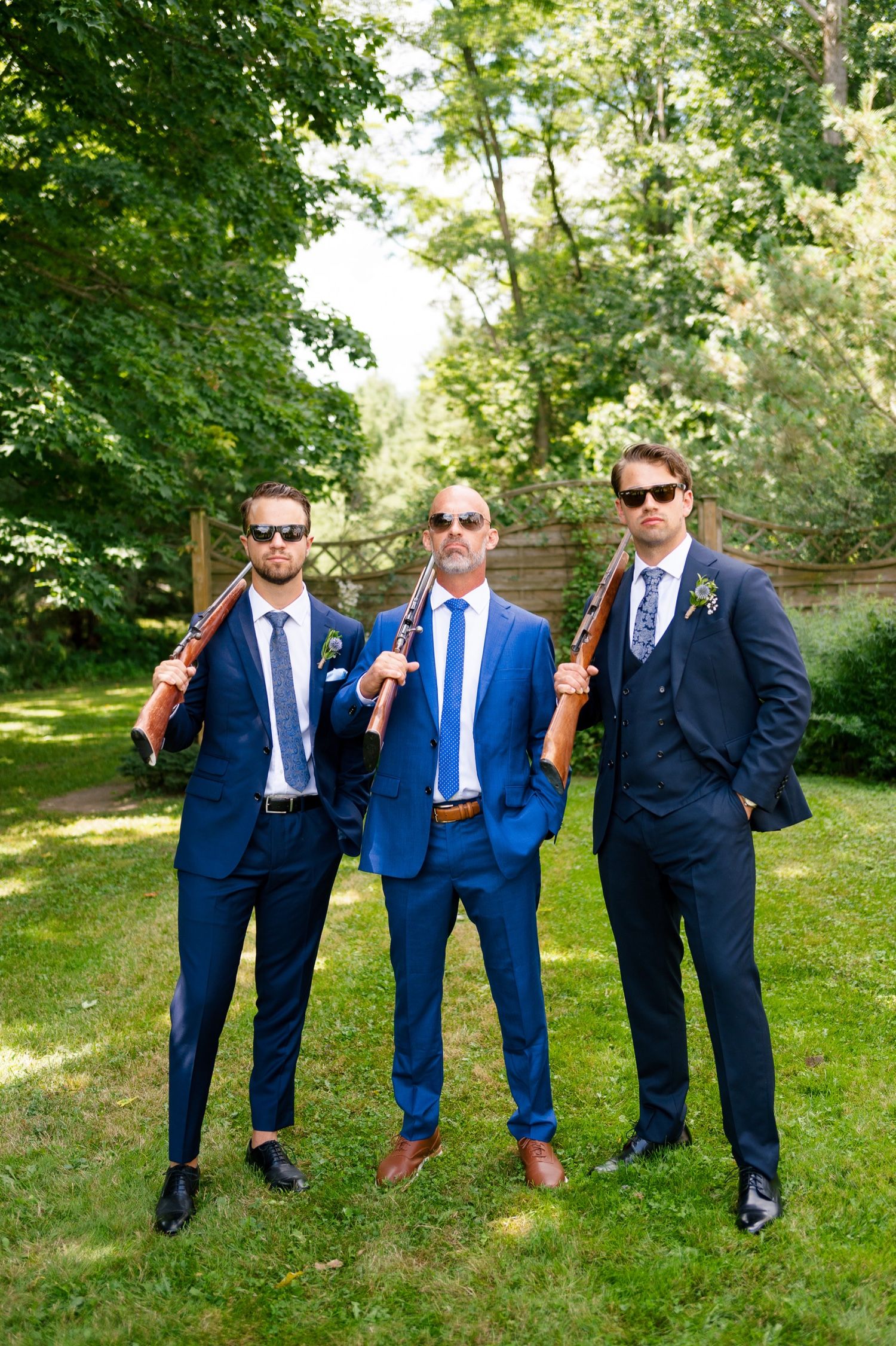 Three groomsmen in sunglasses pose with prop baseball bats in formal suits.