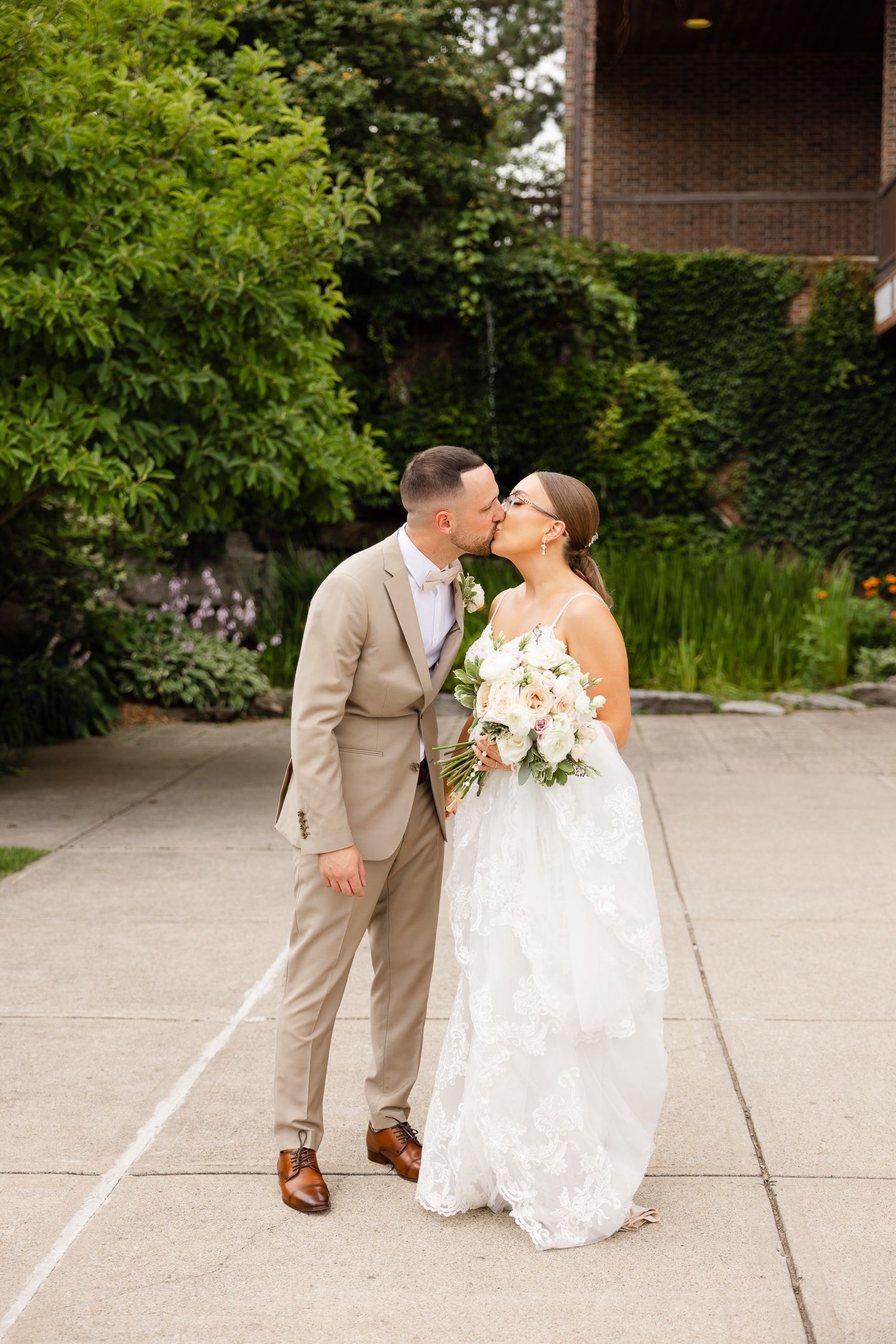 A couple shares a romantic kiss on their wedding day, with the bride in white lace and groom in tan suit.