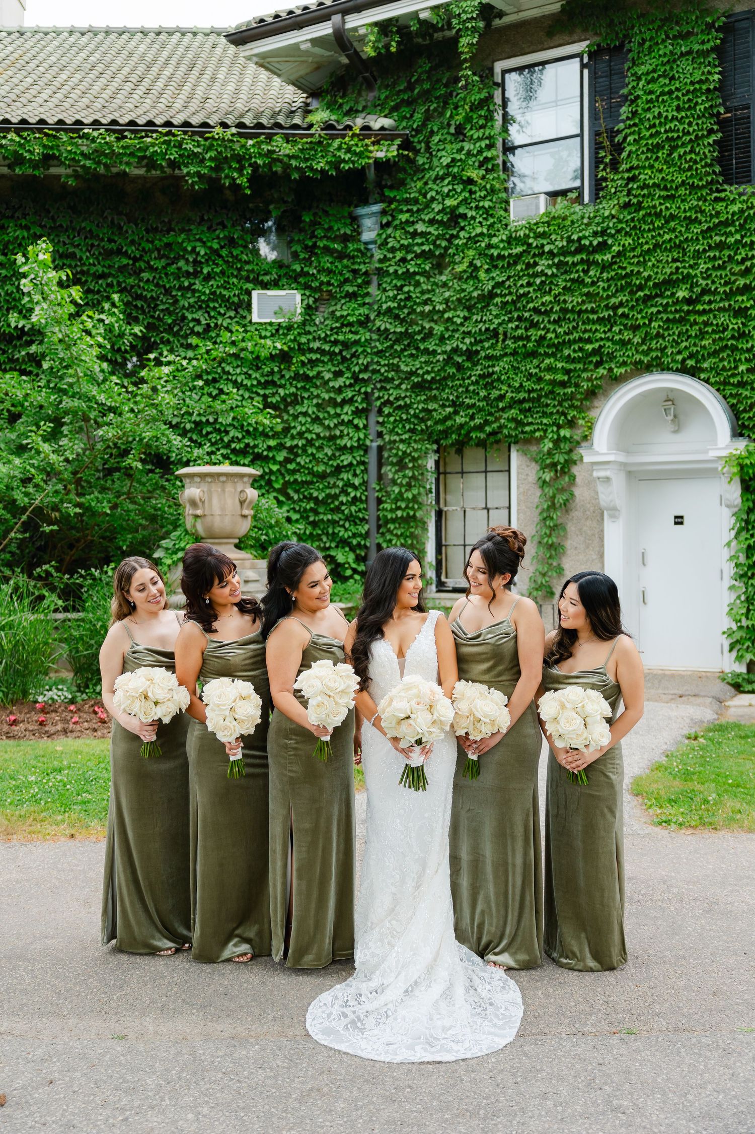 Bridal party in olive green dresses poses with white bouquets against ivy-covered mansion.