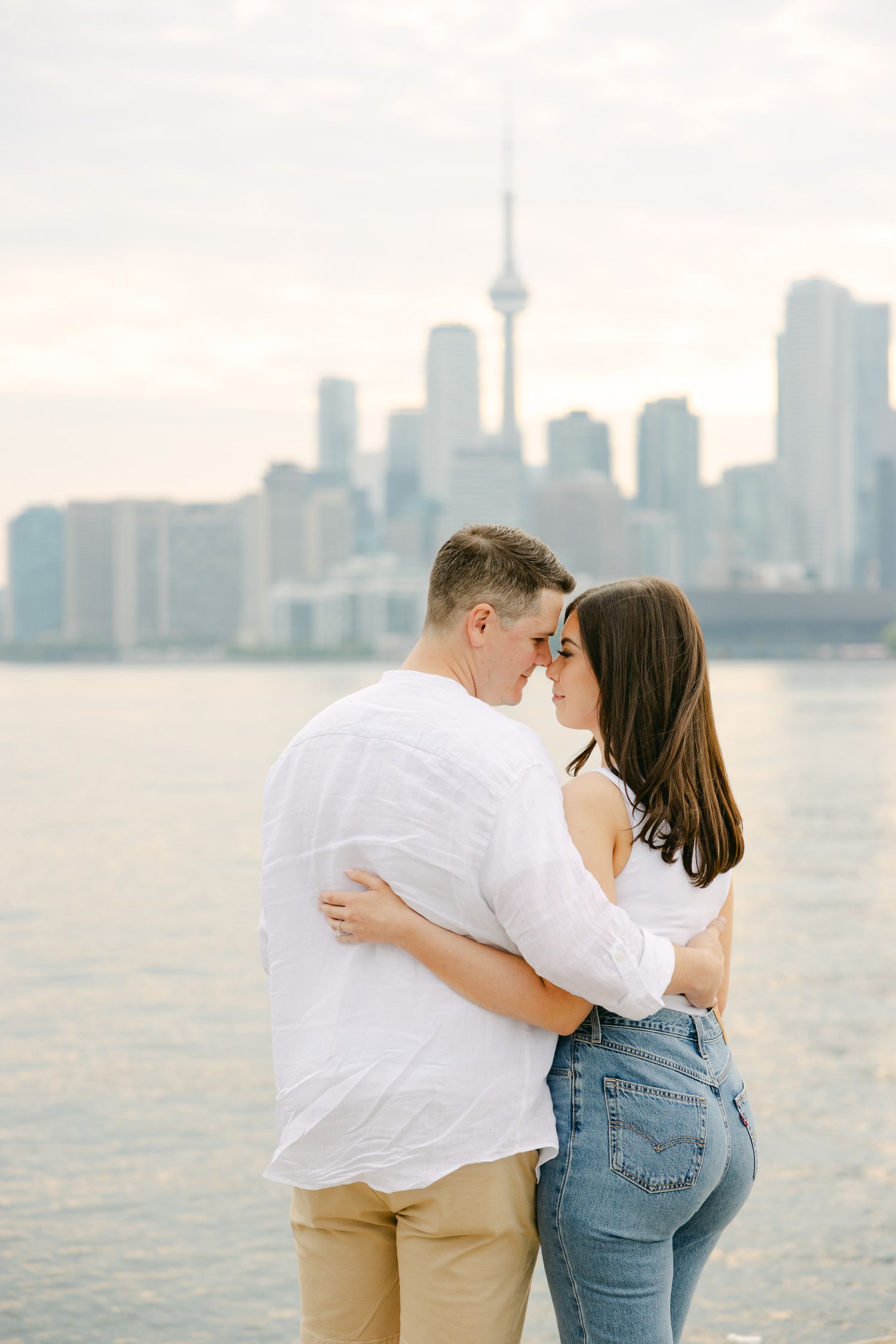 A couple embraces on Toronto's waterfront with the CN Tower and city skyline in the misty background.