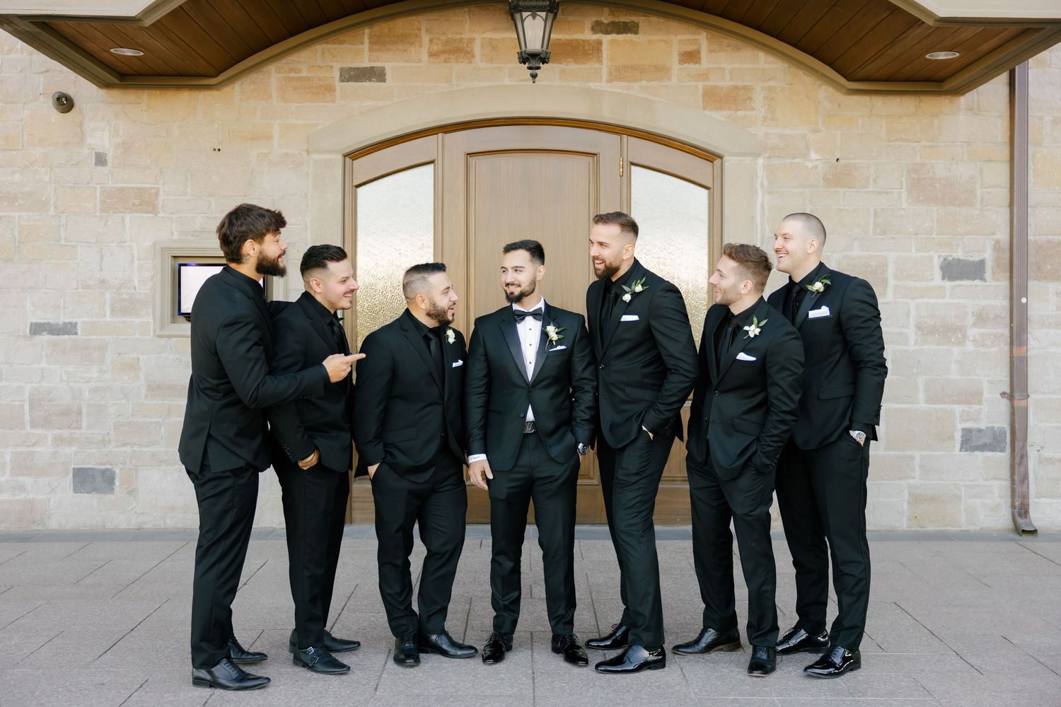 A group of groomsmen share a candid moment while posing under a stone archway.